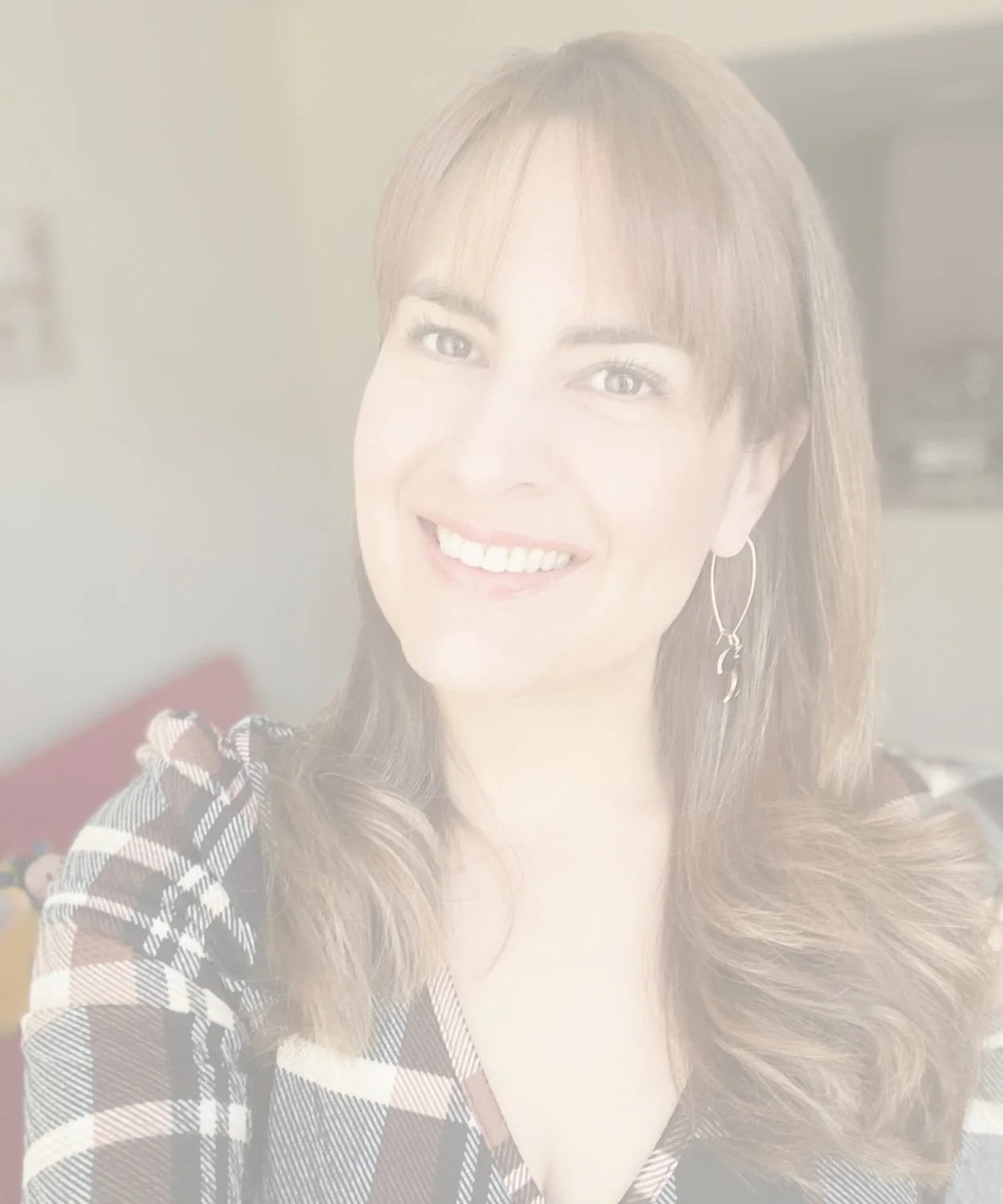 Smiling woman with long, wavy brown hair, wearing hoop earrings and a black, white, and beige checkered top in an indoor setting.