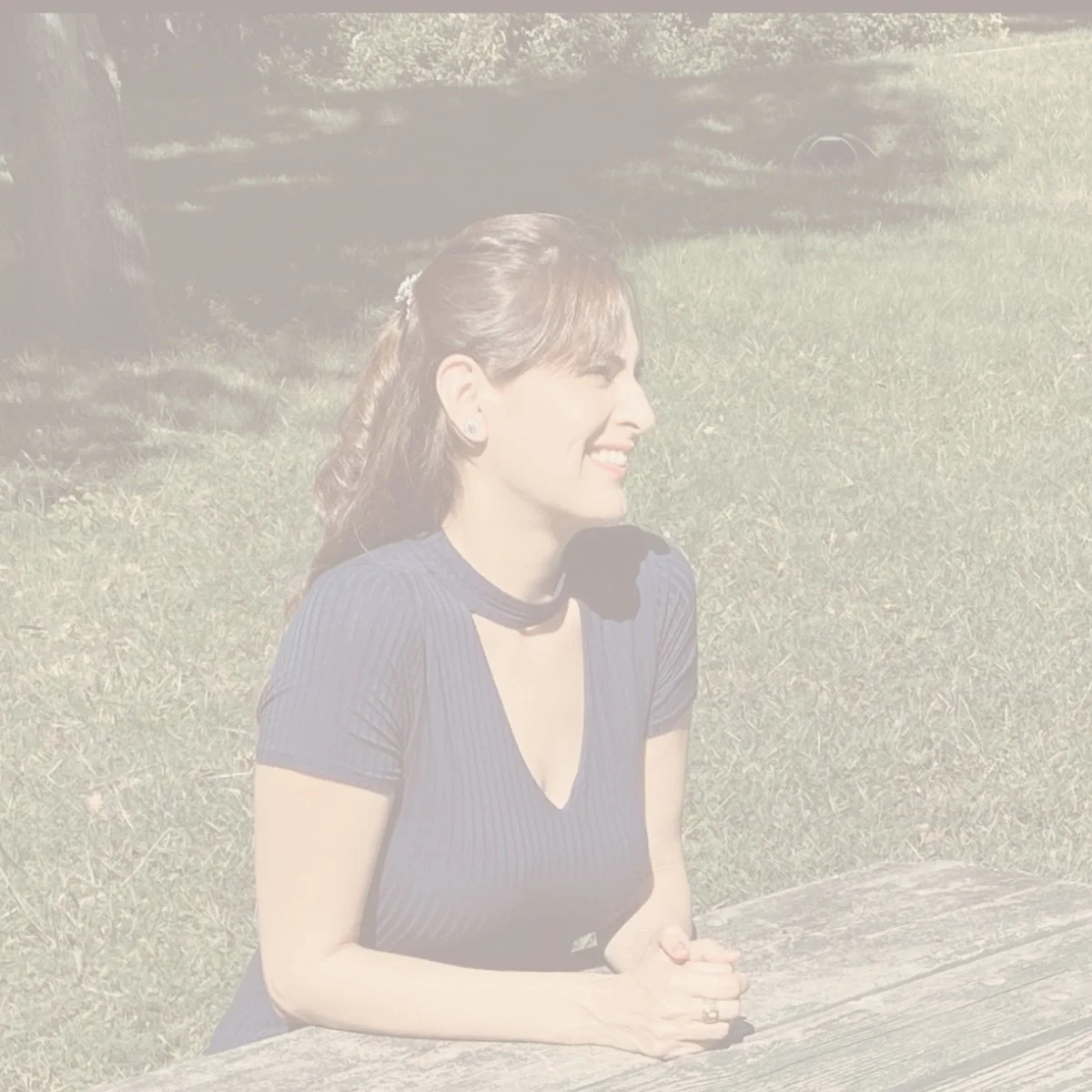 Woman with dark hair tied back, wearing a dark top and pearl earrings, smiling and sitting at a weathered wooden table outdoors on grass, with trees and sunlight in the background.