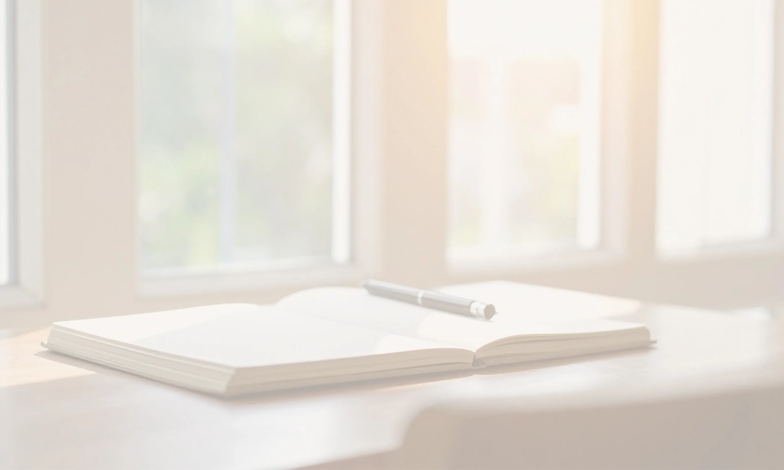 Open notebook with blank pages and a pen on a white desk next to a window with sunlight.