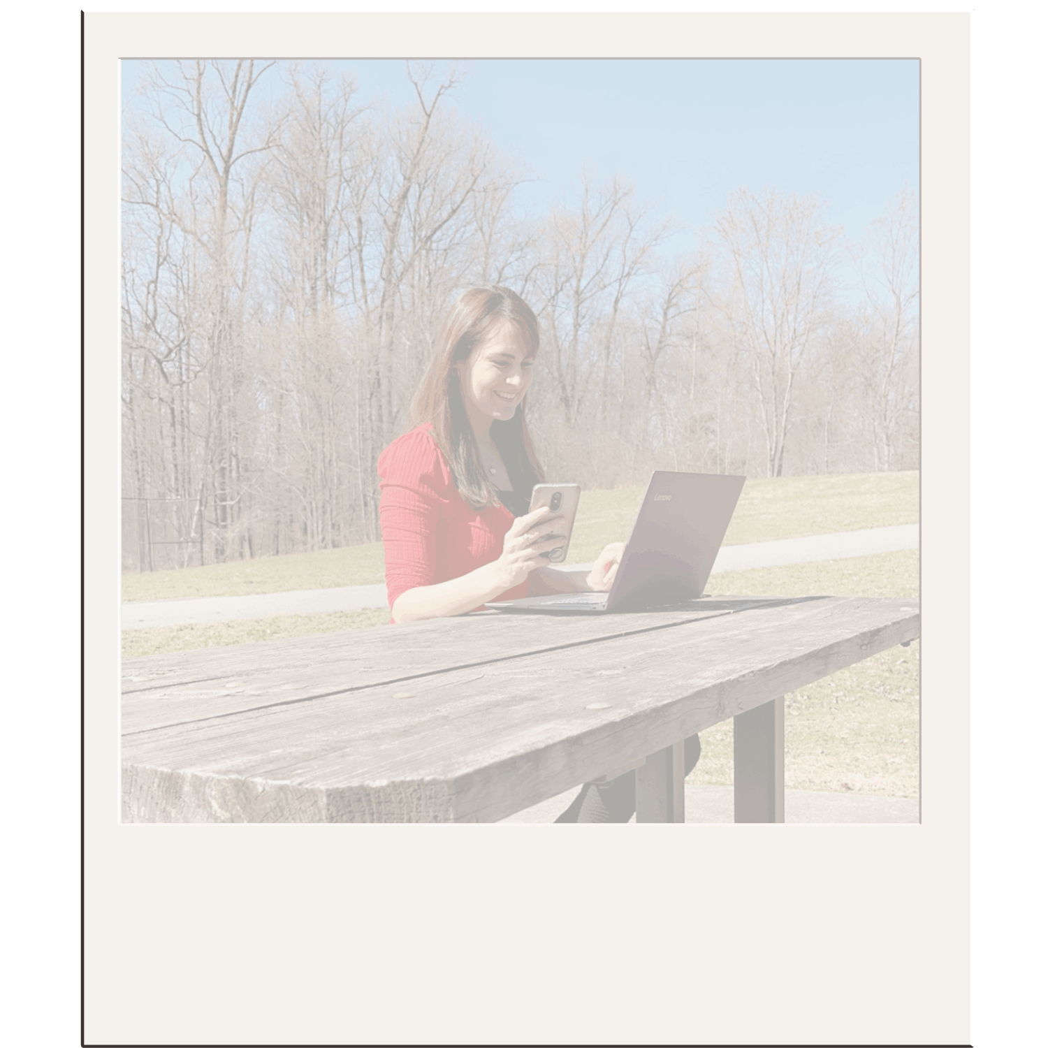 A woman in a red shirt smiling while sitting at a wooden picnic table outdoors, looking at her smartphone with a laptop open in front of her on the table, with trees and a clear sky in the background.