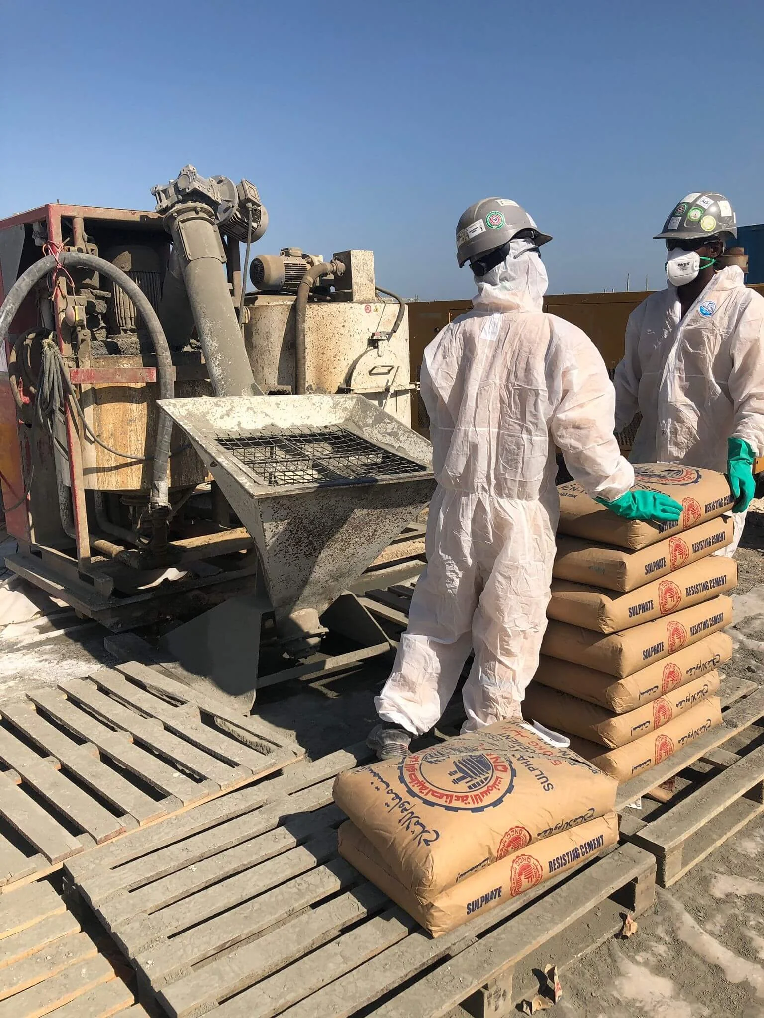 Construction workers in protective gear stack cement bags next to a concrete mixing machine at a job site.