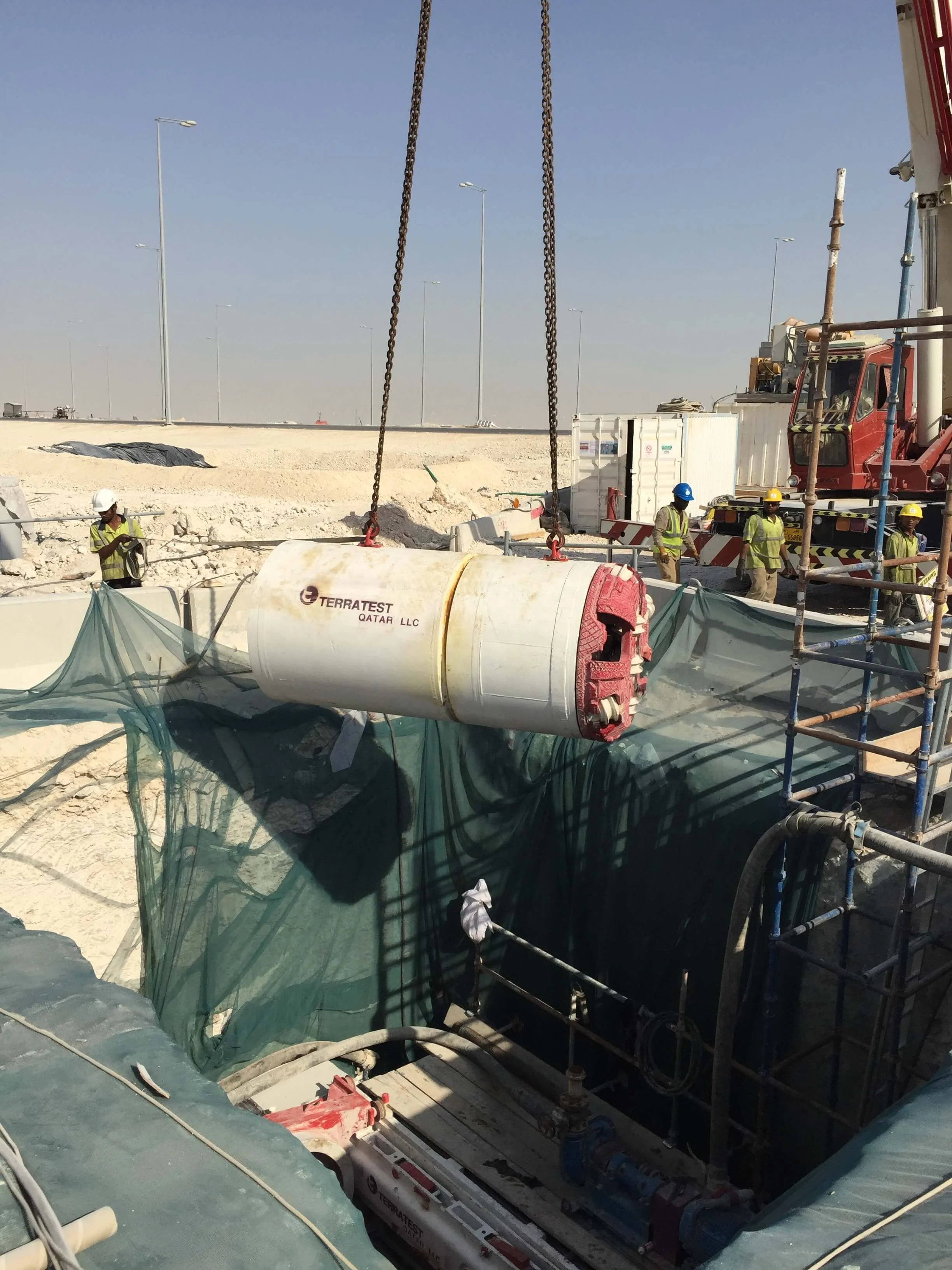 Crane lowers a white microtunneling machine into an excavation trench with construction workers nearby.