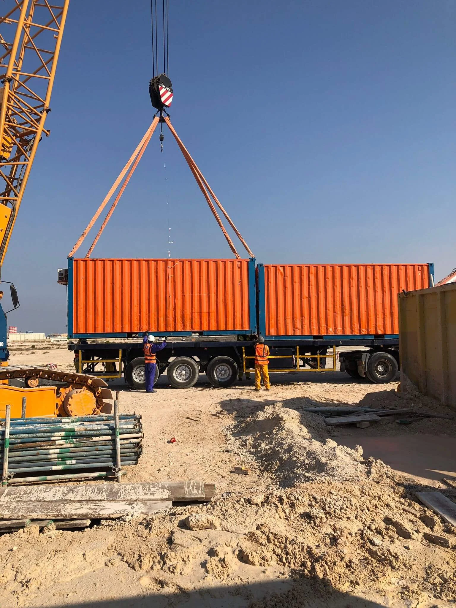 Workers unload orange shipping containers from a truck using a large construction crane.