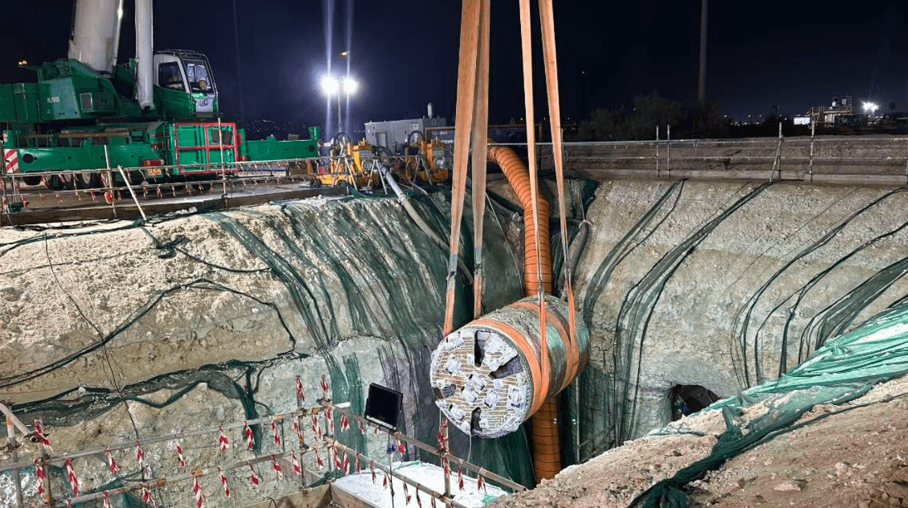 Large green crane lifts tunnel boring machine into deep excavation pit at night. Large-scale construction site.