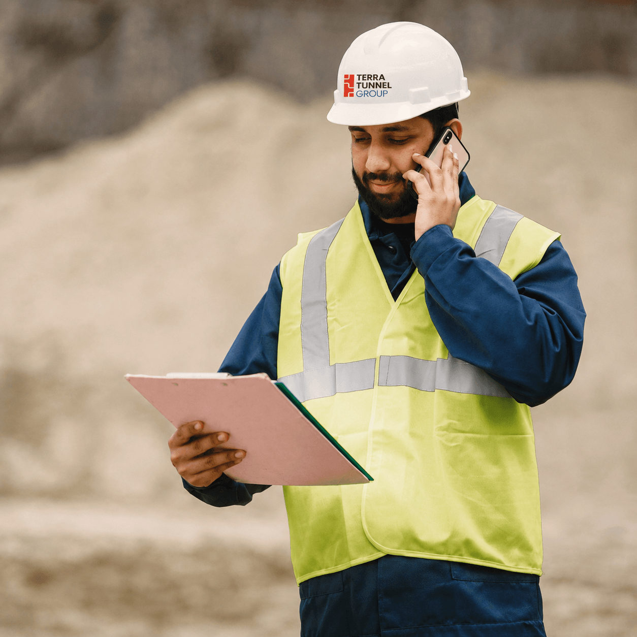 Construction worker on phone wearing safety vest, holding a clipboard.