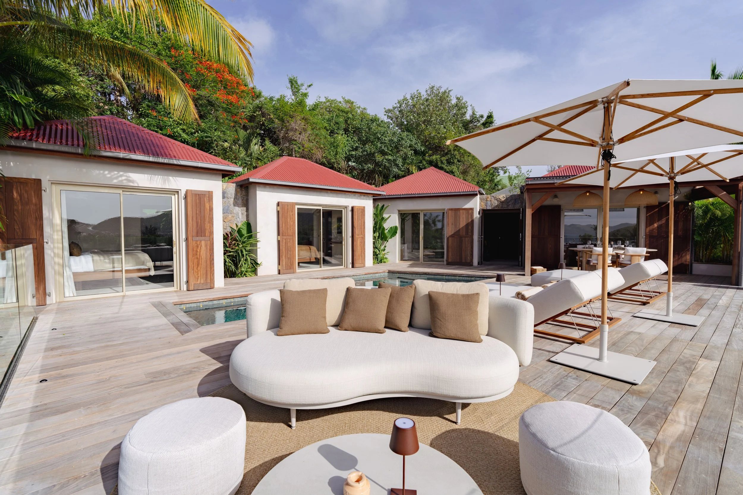 A modern outdoor patio with beige cushioned lounge chairs and matching ottomans, a large white umbrella, a small side table with a brown lamp, and a view of neighboring houses with red roofs and large windows surrounded by lush green trees.