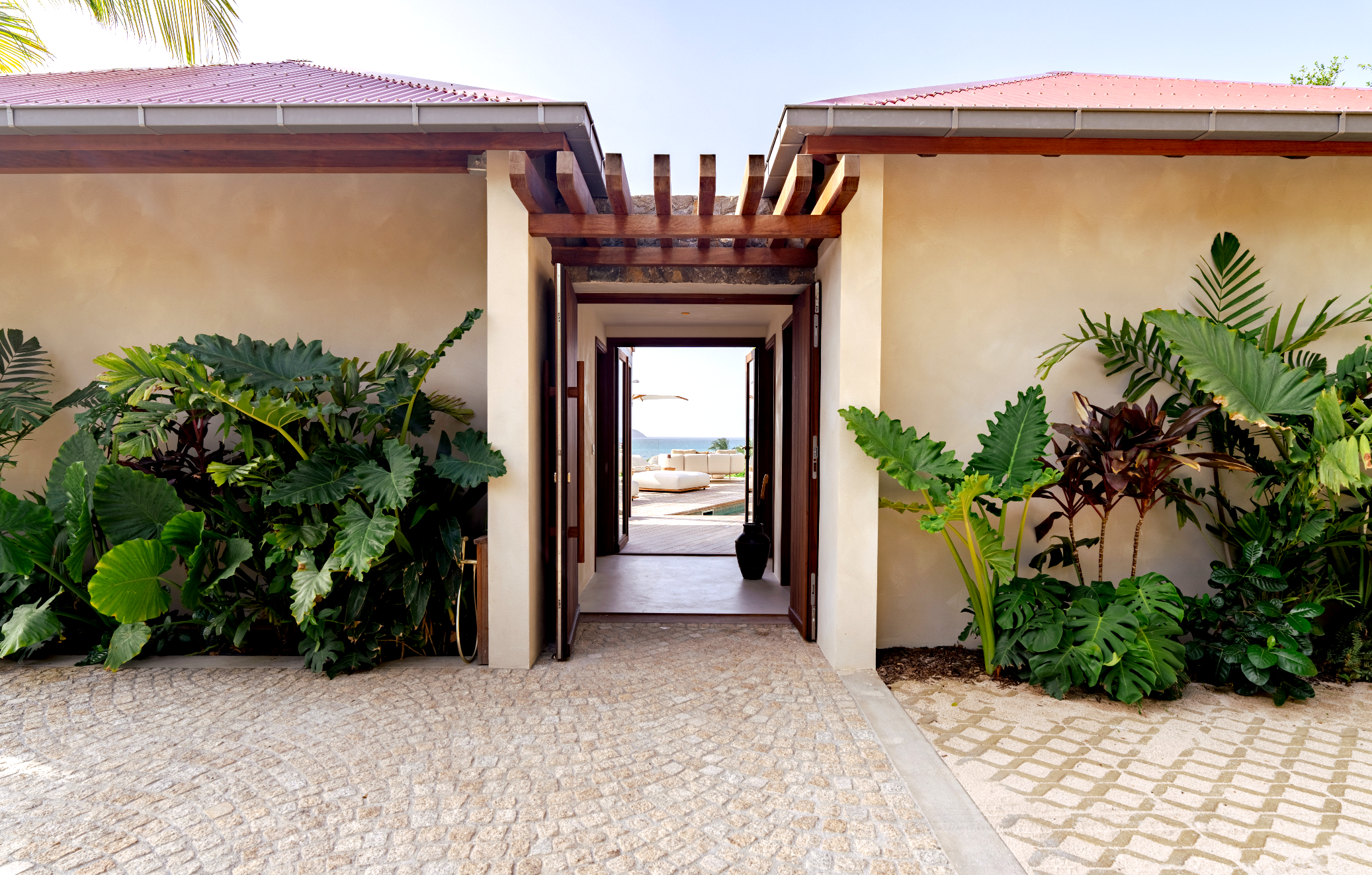 View through open door leading to outdoor patio with white seating, overlooking ocean, framed by lush green tropical plants and an arched stone path.