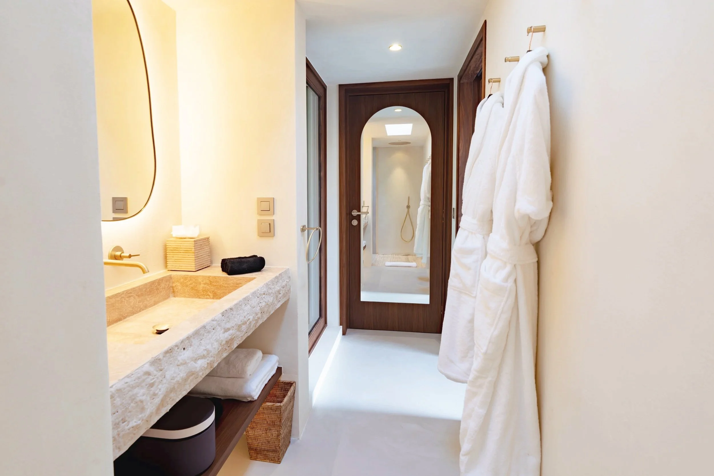 A bathroom with a beige stone countertop, two towels, a mirror, and a wooden door leading to a shower area with a glass enclosure and a handheld showerhead.