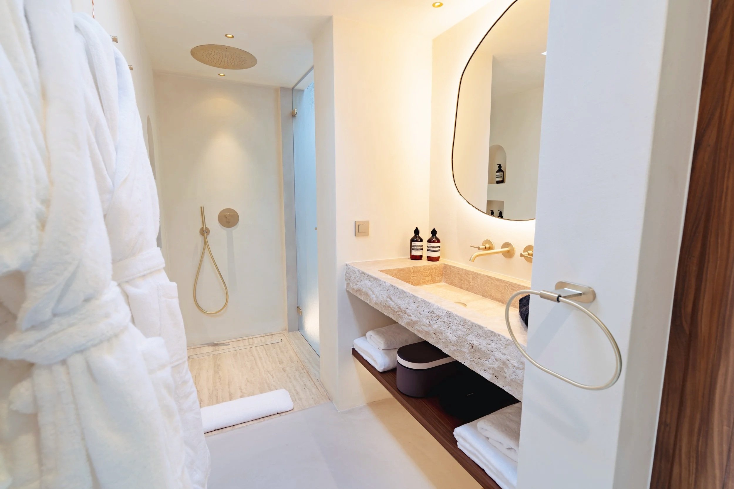A modern, minimalist bathroom with a large round mirror, a stone sink with gold fixtures, and a wall-mounted shower head. Towels are neatly folded on a shelf below the sink, and two bottles of liquid soap or lotion are placed on the countertop.