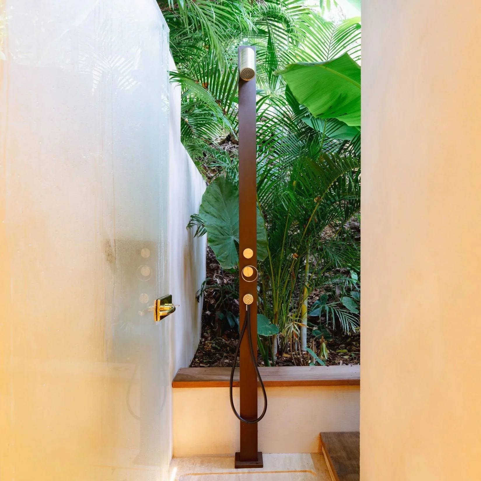 A standing shower head with a brown wooden stand, positioned in front of a view of lush green tropical plants outside a window, with a yellow wall on either side.