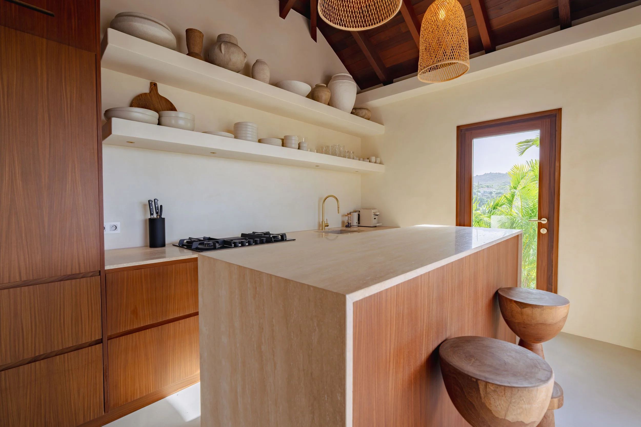 Modern kitchen with white countertops, wooden cabinets, open shelving with pottery, window showing greenery outside, and two wooden stools by the counter.