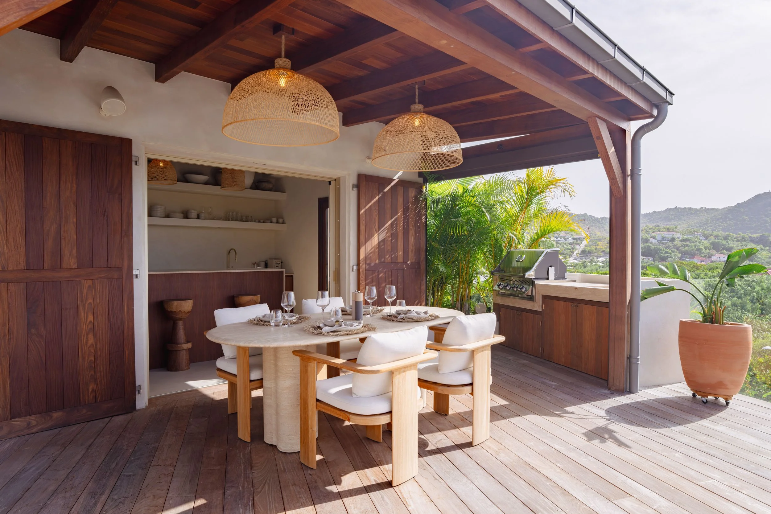 An outdoor patio area with a dining table set for four, surrounded by white cushioned chairs, wooden accents, ceiling pendant lights, potted plants, and a view of distant hills.