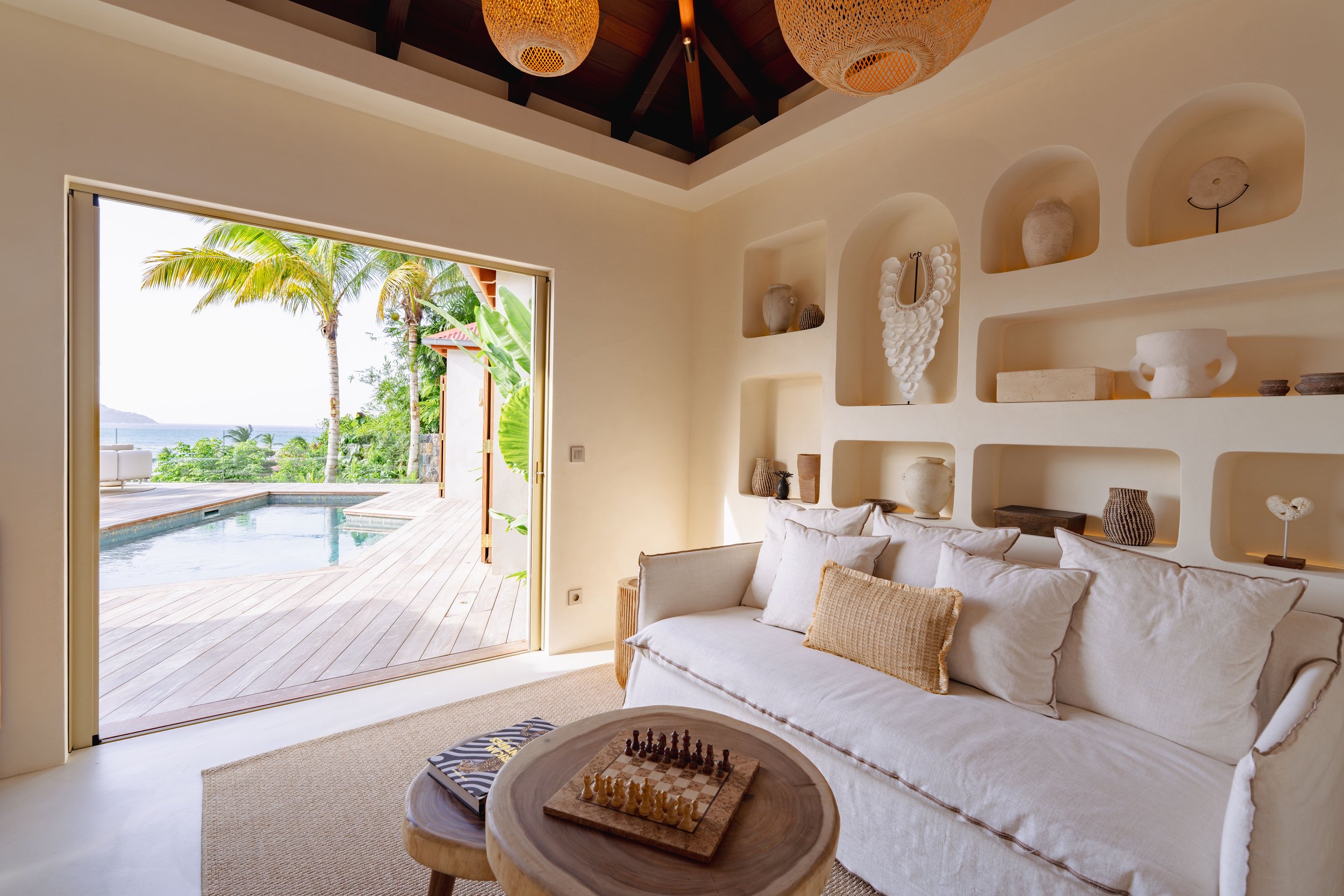 A cozy living room with a white couch and cushions, large open doorway leading to a wooden deck with a pool, overlooking palm trees and the ocean, and decorative shelves with vases and ornaments.