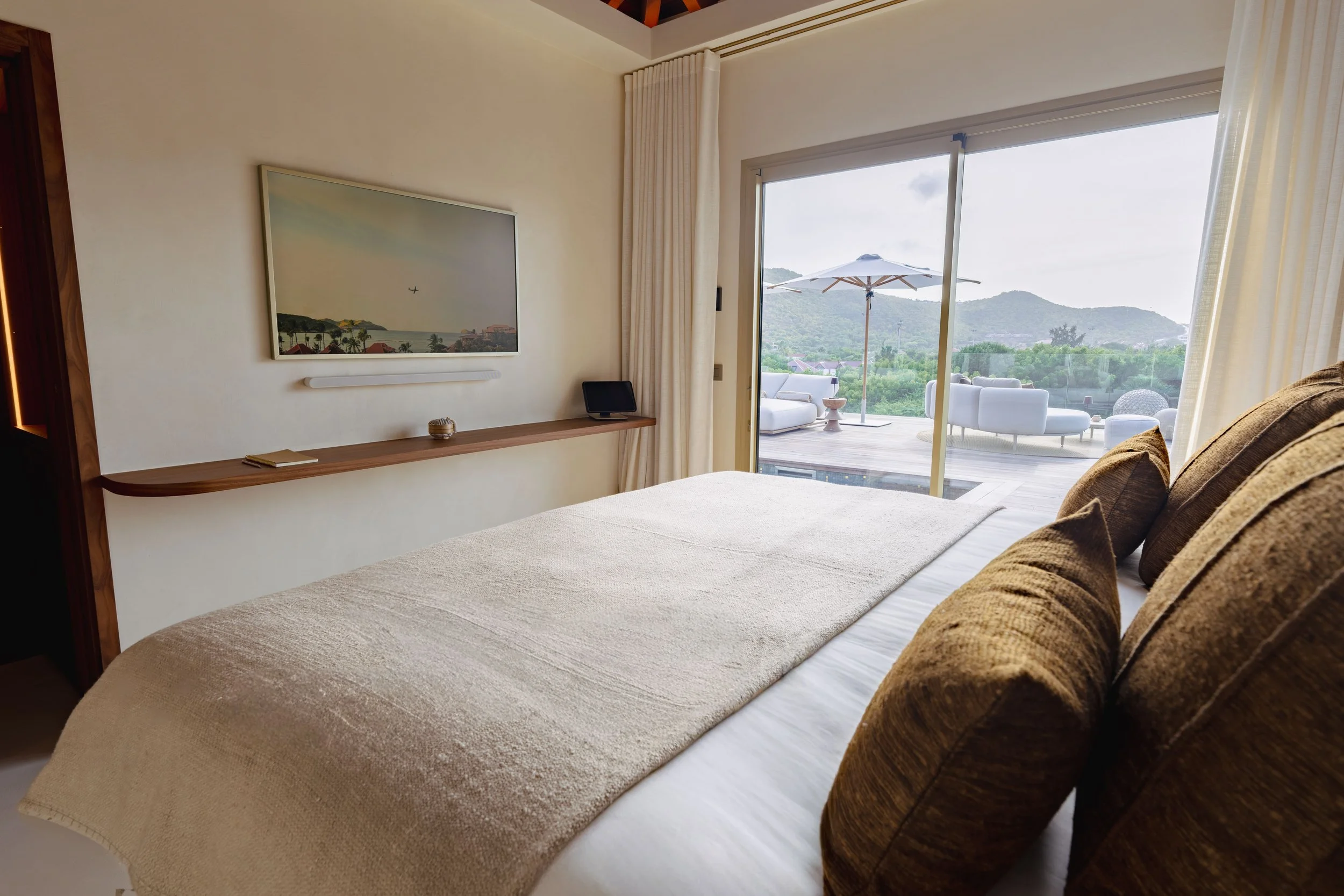 A bedroom with a bed covered in a beige bedspread and brown pillows, a wall-mounted flat-screen TV, a wooden shelf with a small notebook, a balcony with outdoor furniture, including white chairs and a large umbrella, overlooking a green landscape wit