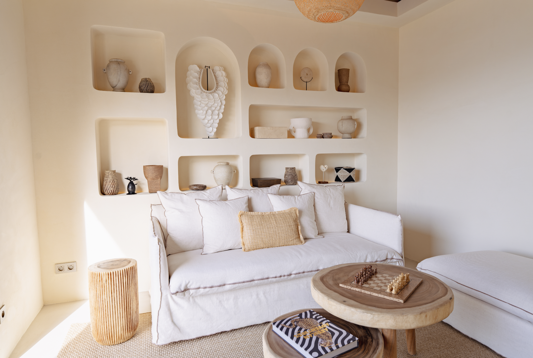 Living room with white sofa, beige pillows, round wooden coffee table with chess and a magazine, wall with niche shelves displaying vases and decorative objects, cream walls, wicker side table, woven hanging lampshade.