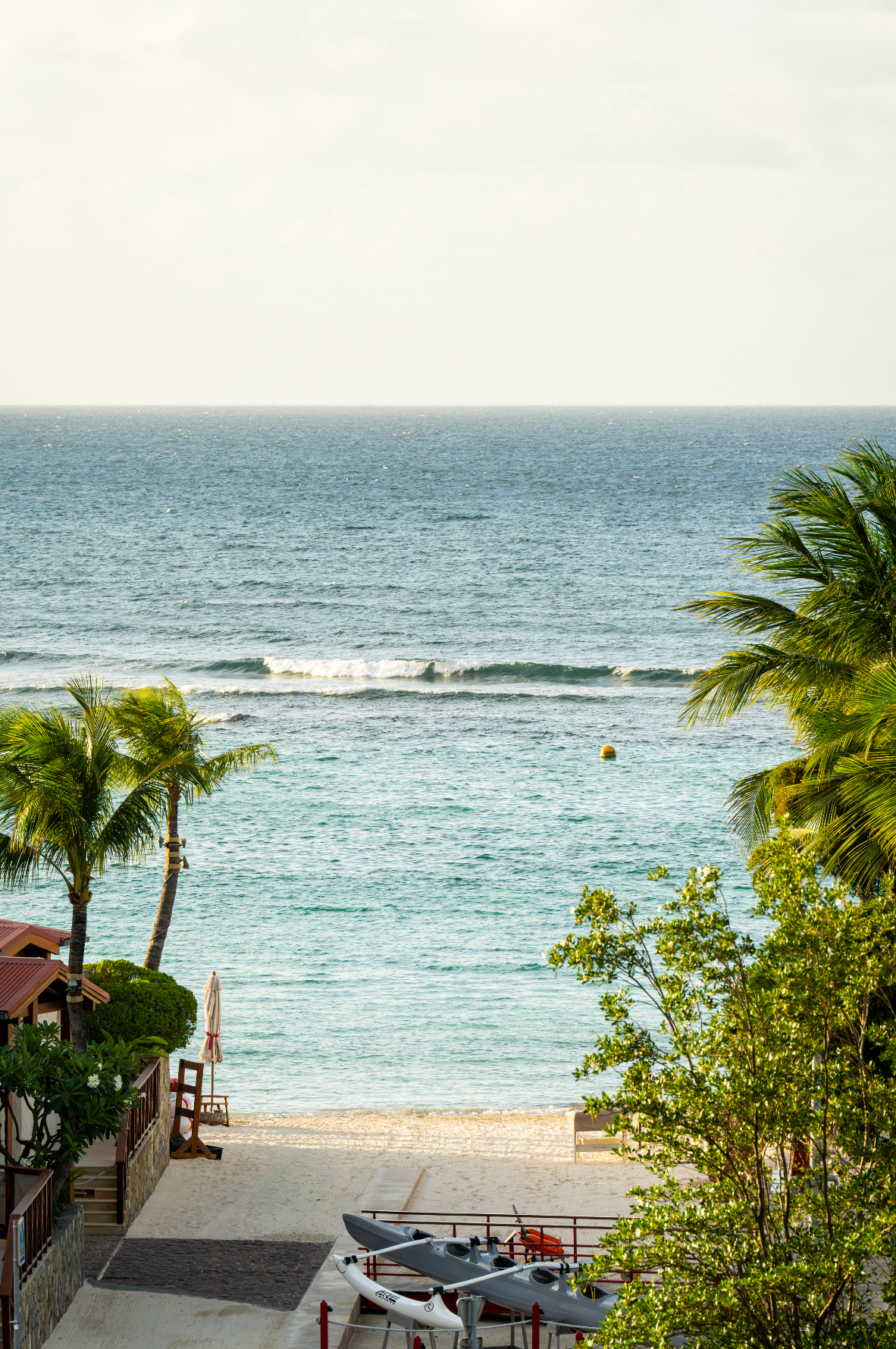 View of a beach with palm trees, a boat, and the ocean with gentle waves and a buoy, under a hazy sky.