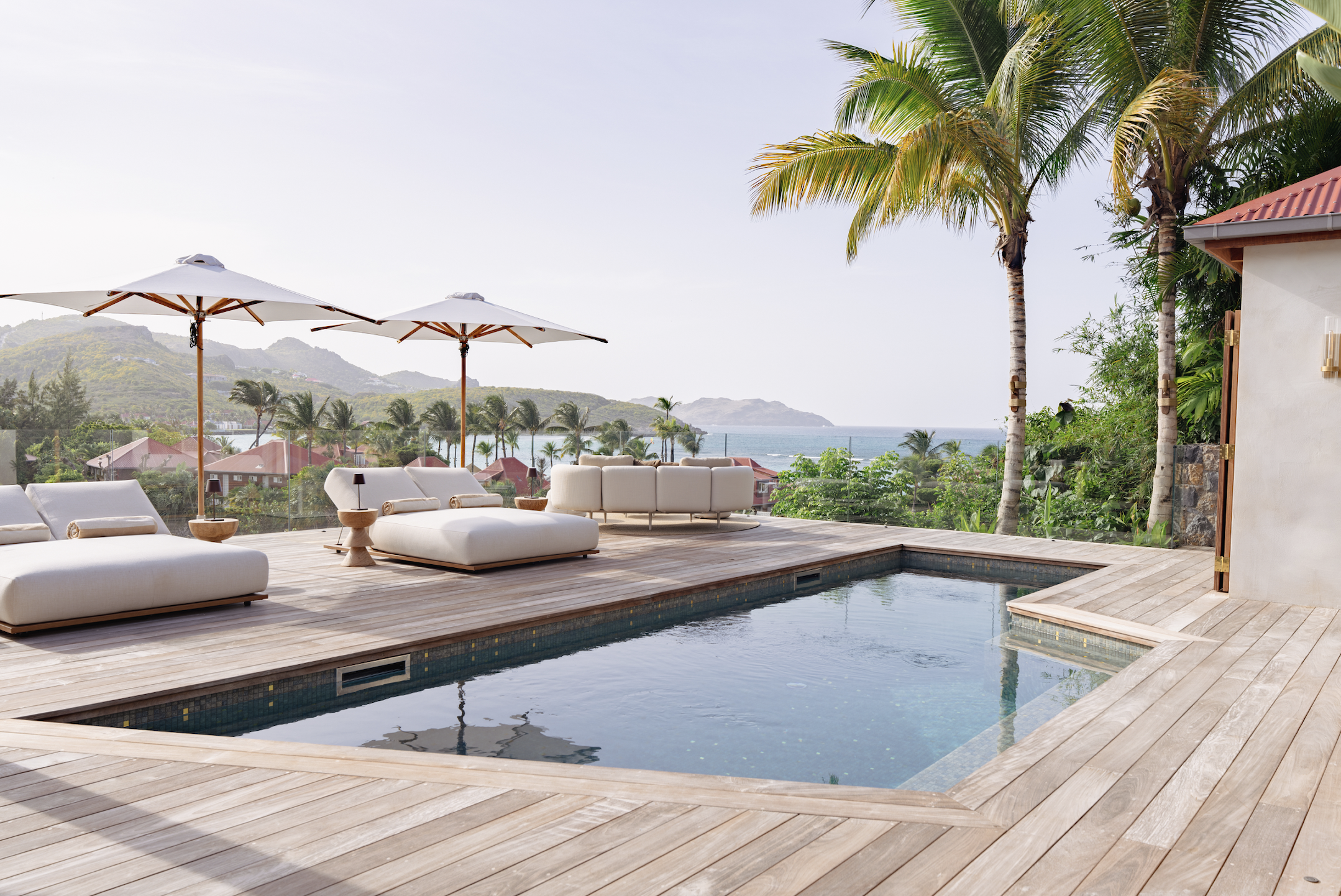 A luxurious outdoor pool area with white cushioned loungers, white umbrellas, and a view of palm trees, hills, and the ocean in the background.