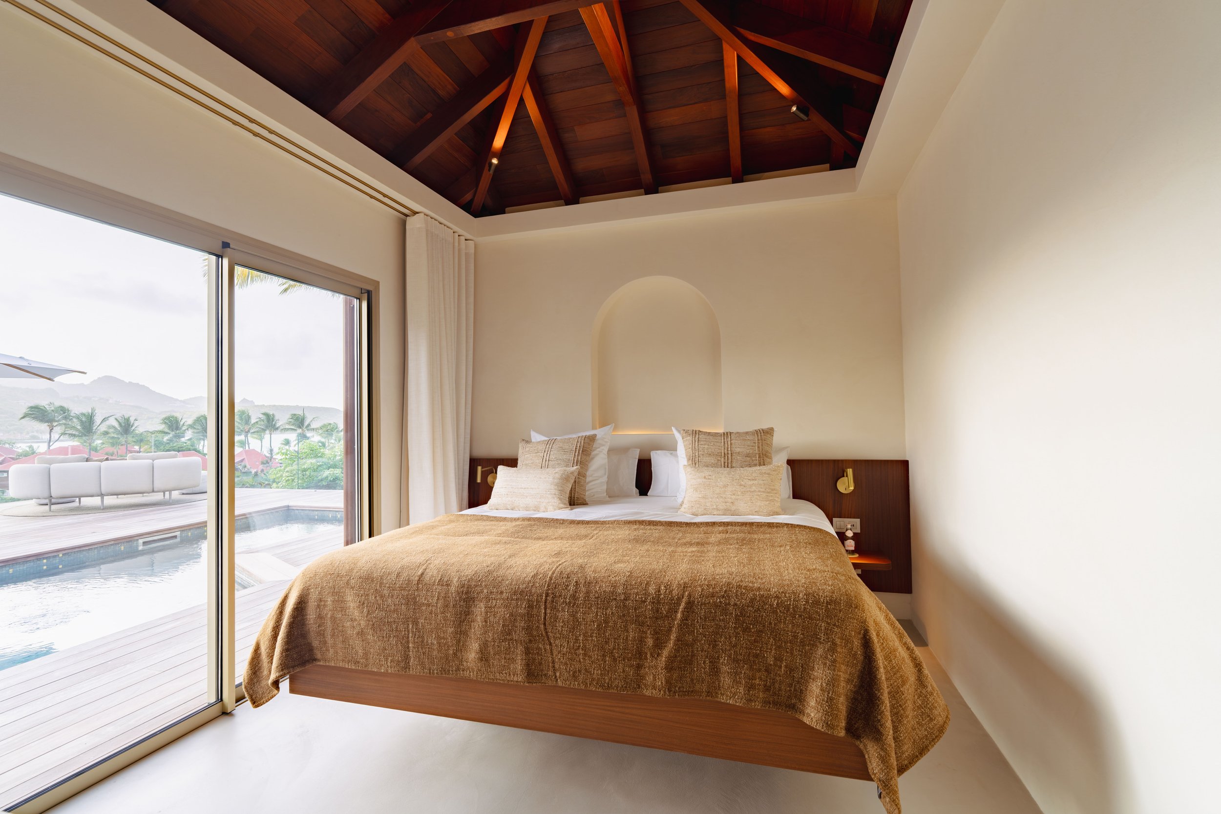 A bedroom with a large bed covered in a brown blanket, beige pillows, and a wooden headboard. The room has a high, vaulted wooden ceiling with spotlights. Large sliding glass doors open to a balcony with outdoor patio furniture, a pool, and a view of