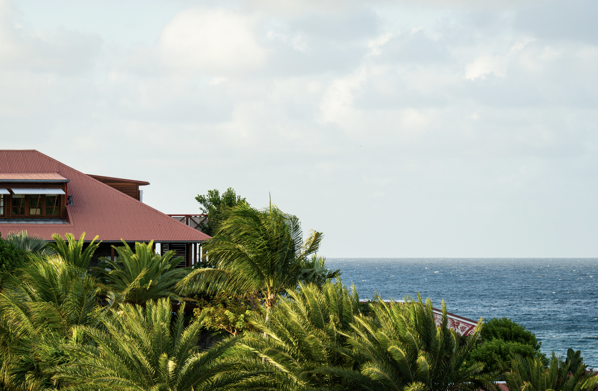 A coastal house with a red roof surrounded by lush green tropical plants, overlooking the ocean under a cloudy sky.