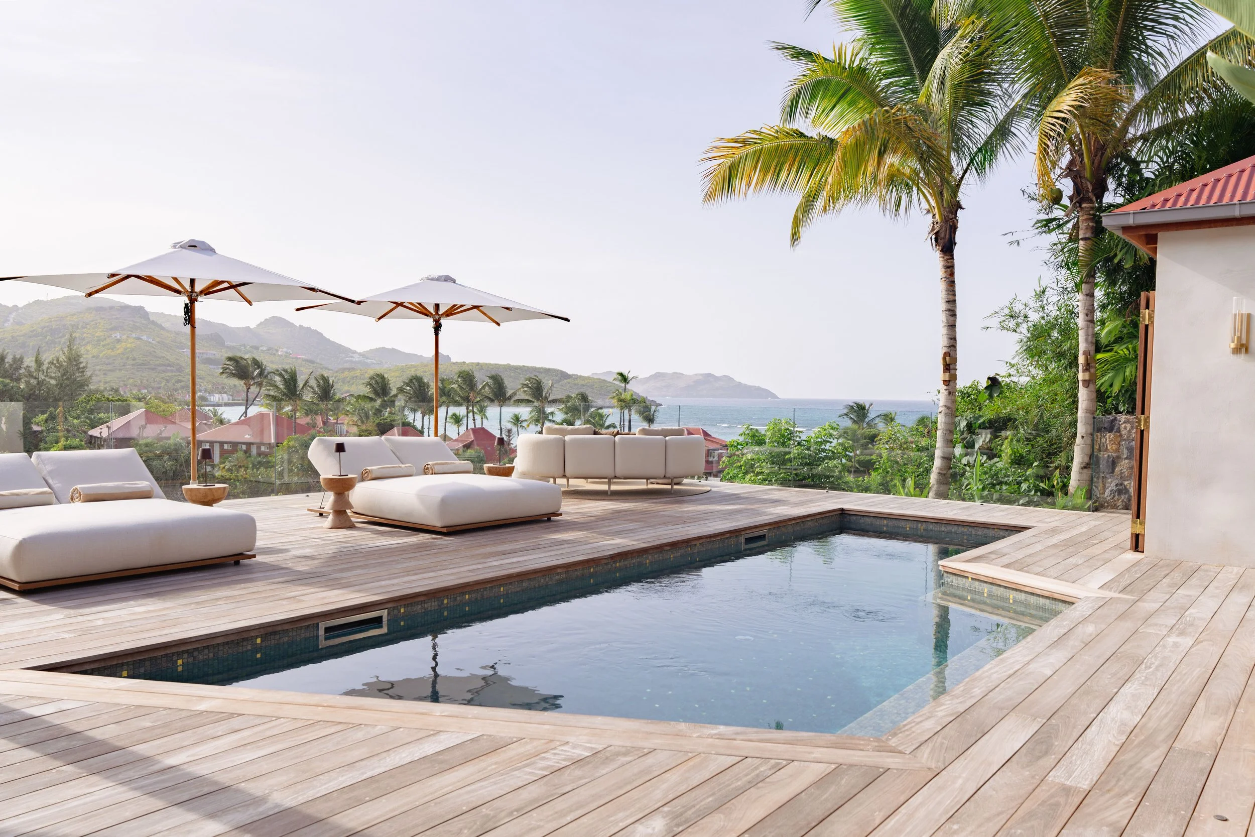Luxury outdoor patio overlooking the ocean with pool, white lounge chairs, umbrellas, palm trees, and hillside houses in the distance.