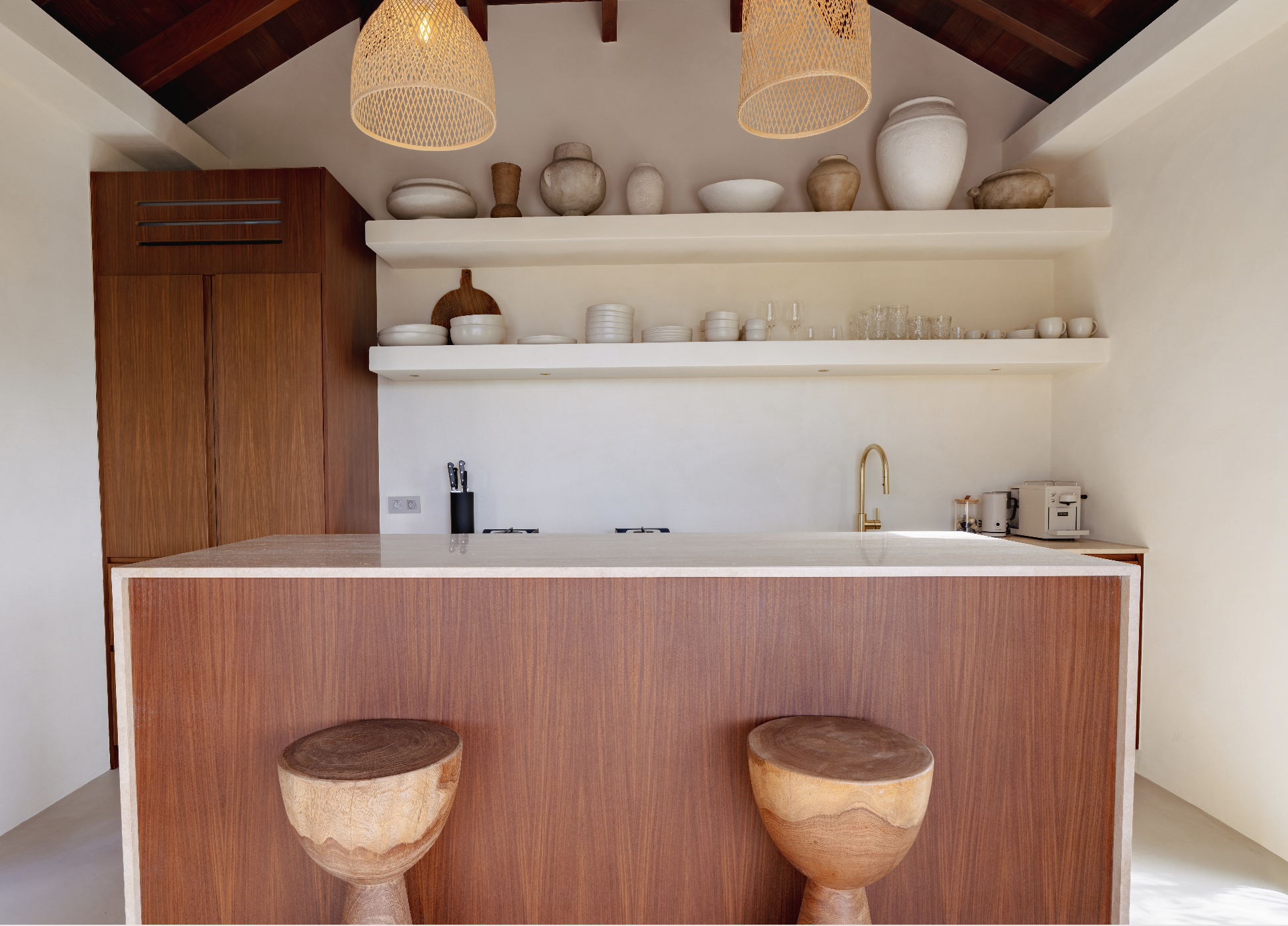 Interior of a modern kitchen with open shelves holding white and beige pottery, a wooden cabinet, and a kitchen island with two wooden stools. The ceiling has exposed dark wooden beams and two woven pendant lights.