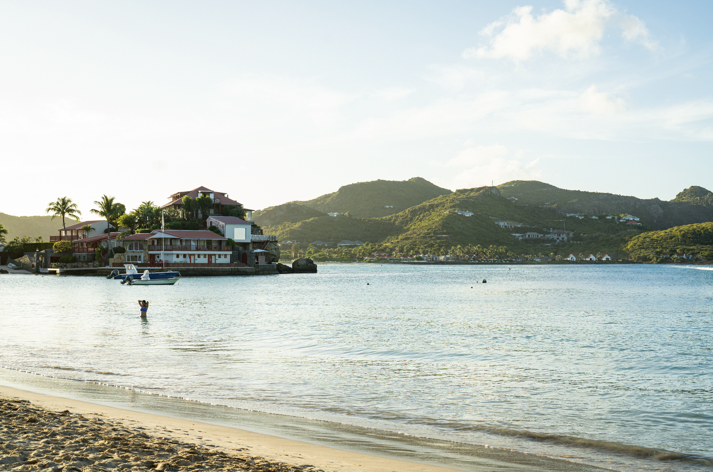 Beach with calm water, a person wading in the water, houses and yacht near the shoreline, green hills in the background, and blue sky with some clouds.