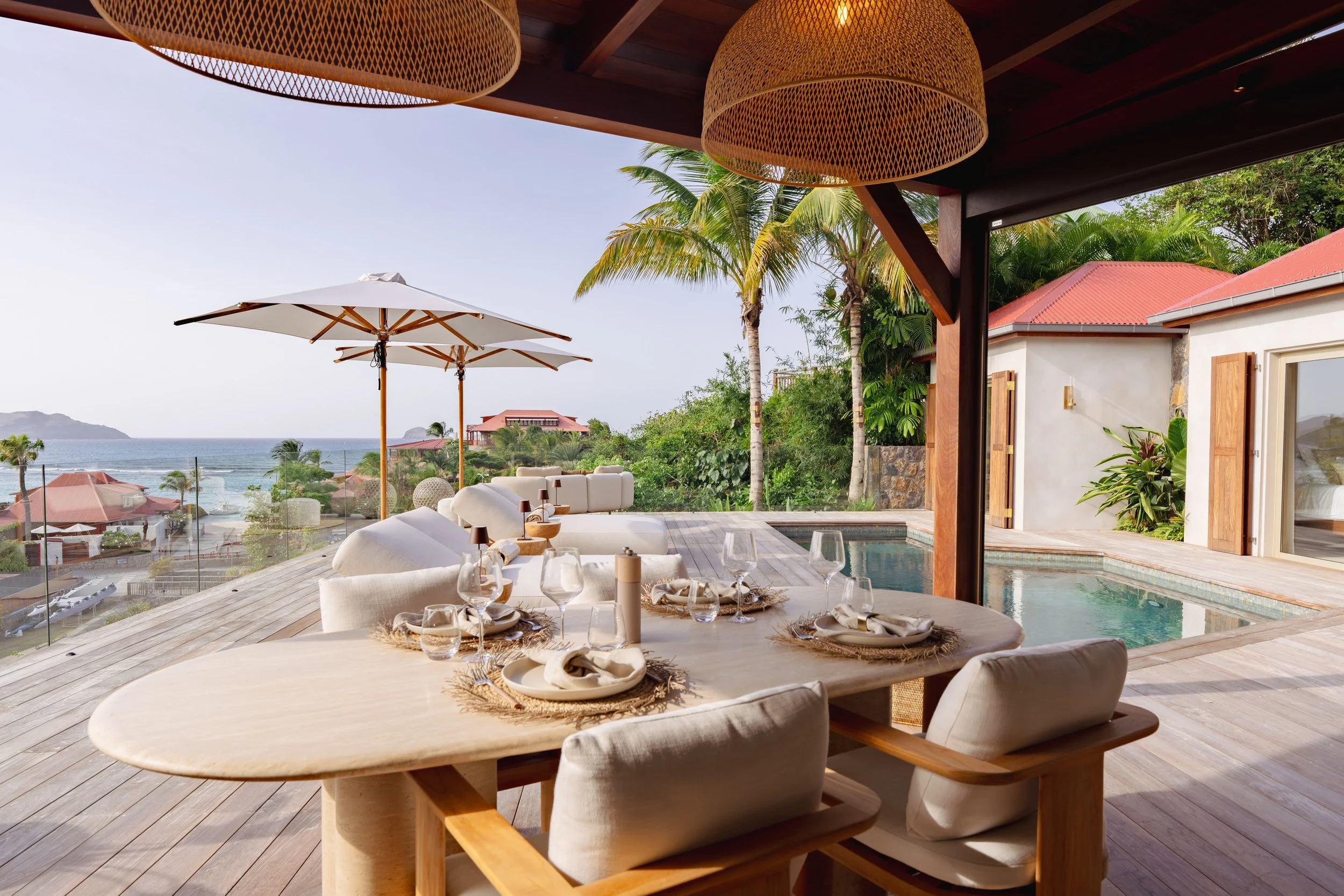 Outdoor patio with a large dining table set with glassware, plates, and napkins, overlooking a scenic ocean view with palm trees and beach umbrellas.