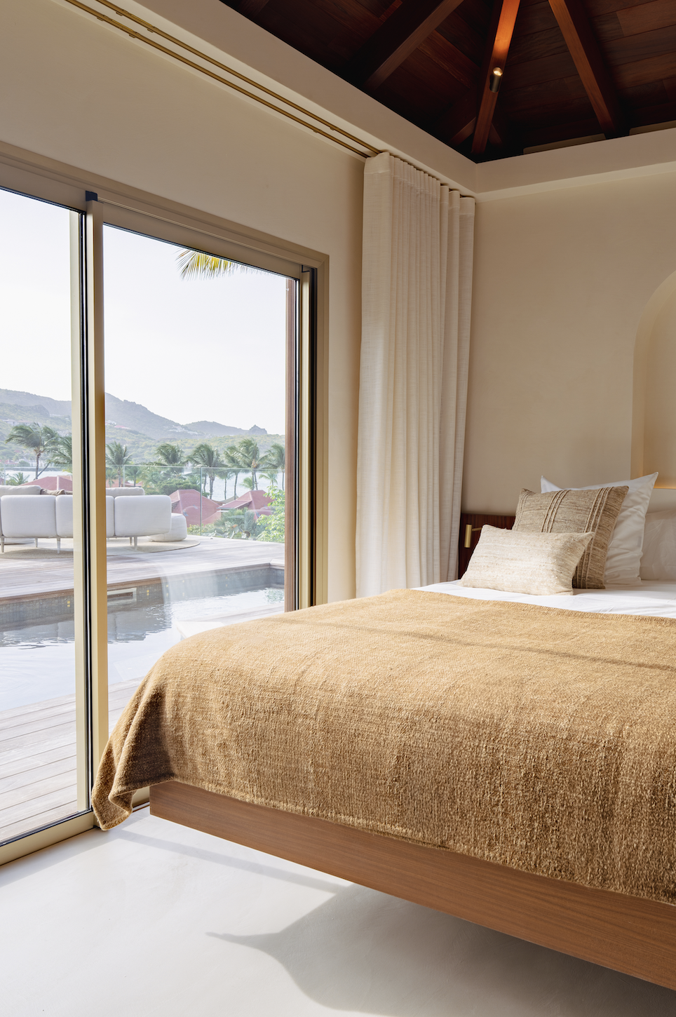 A bedroom with a bed covered in a beige blanket and pillows, next to a sliding glass door opening to a balcony with outdoor seating and a view of palm trees, water, and mountains.