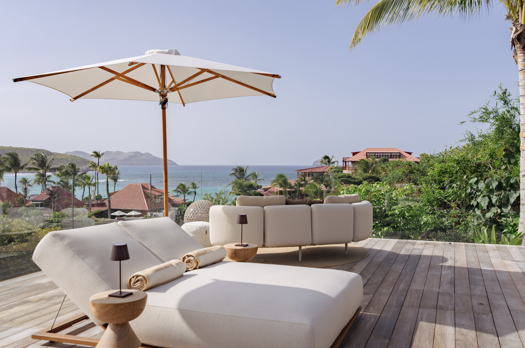View of an outdoor patio with lounge chairs, a large umbrella, and lush greenery overlooking an ocean with mountains in the distance.