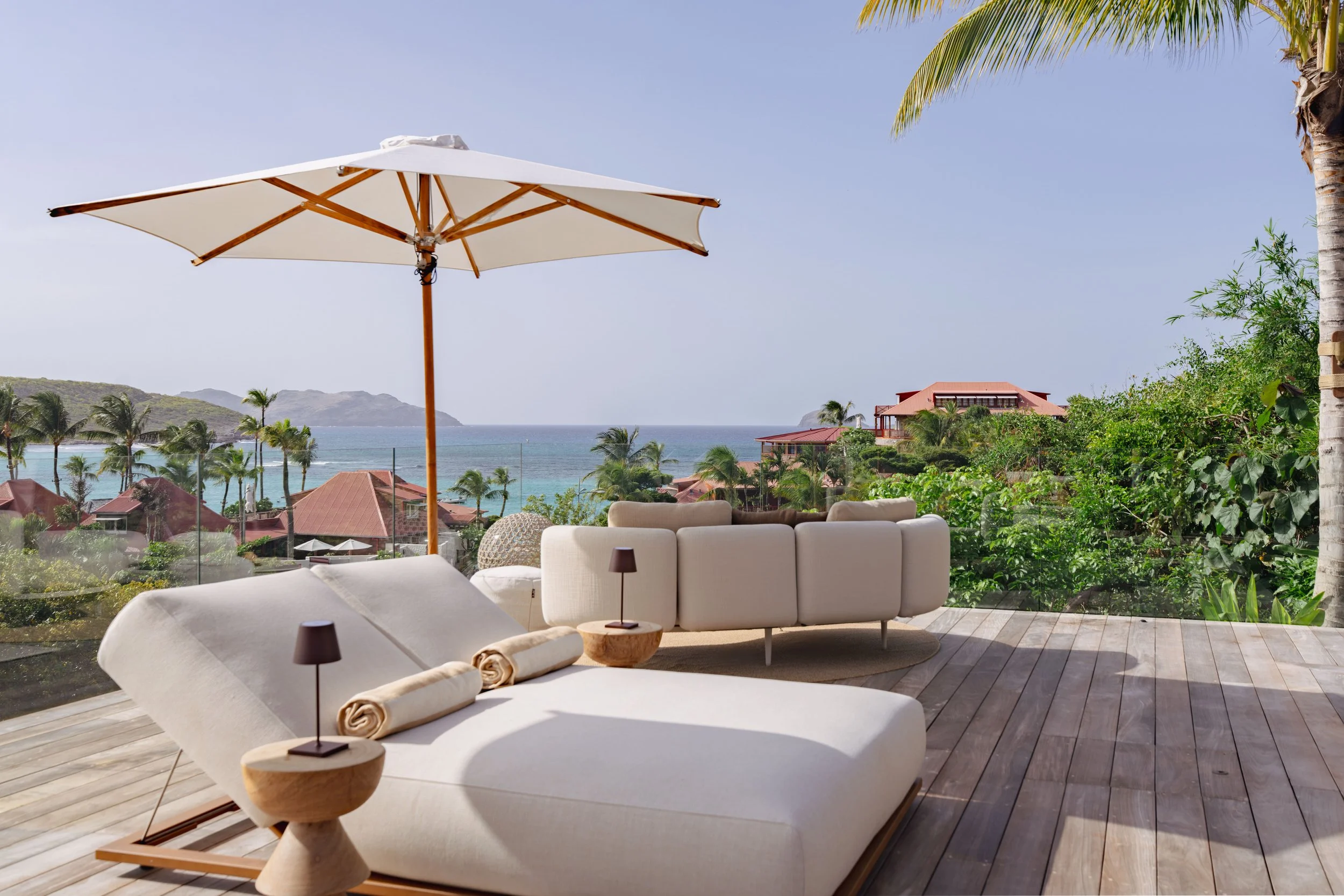 Outdoor patio with white lounge chairs, rolled towels, small tables, large umbrella, and scenic view of beach, palm trees, and mountains in the background.