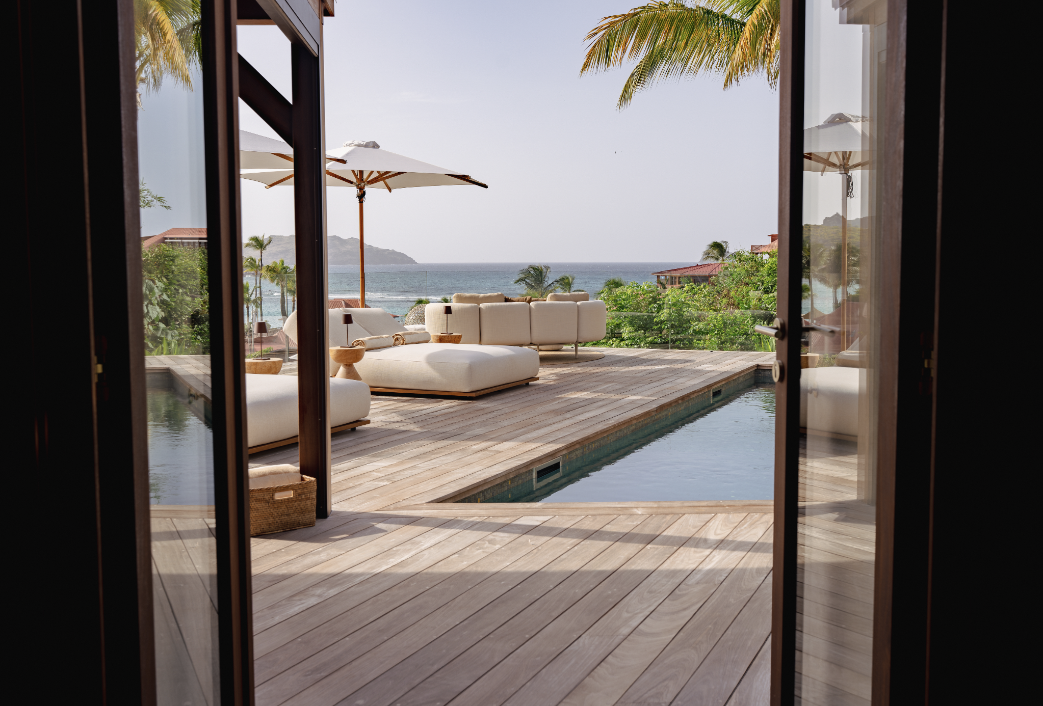 View of a tropical outdoor patio with white outdoor furniture, a large umbrella, palm trees, and a view of the ocean and distant hills, seen through an open doorway.