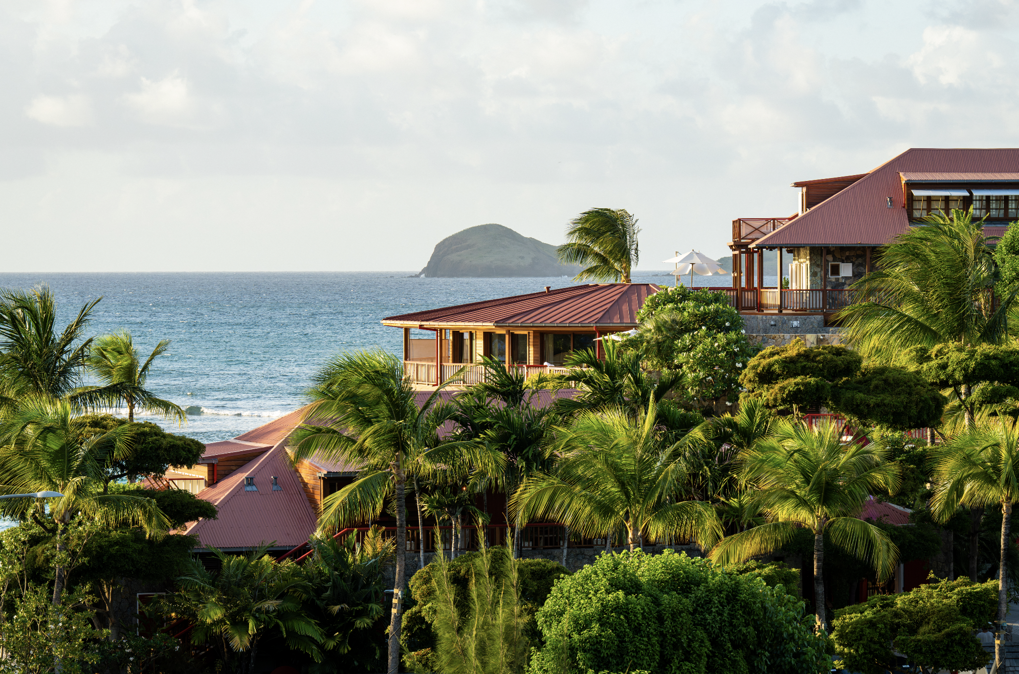 Tropical resort with red roof buildings amid lush green palm trees overlooking the ocean with a small island in the background.