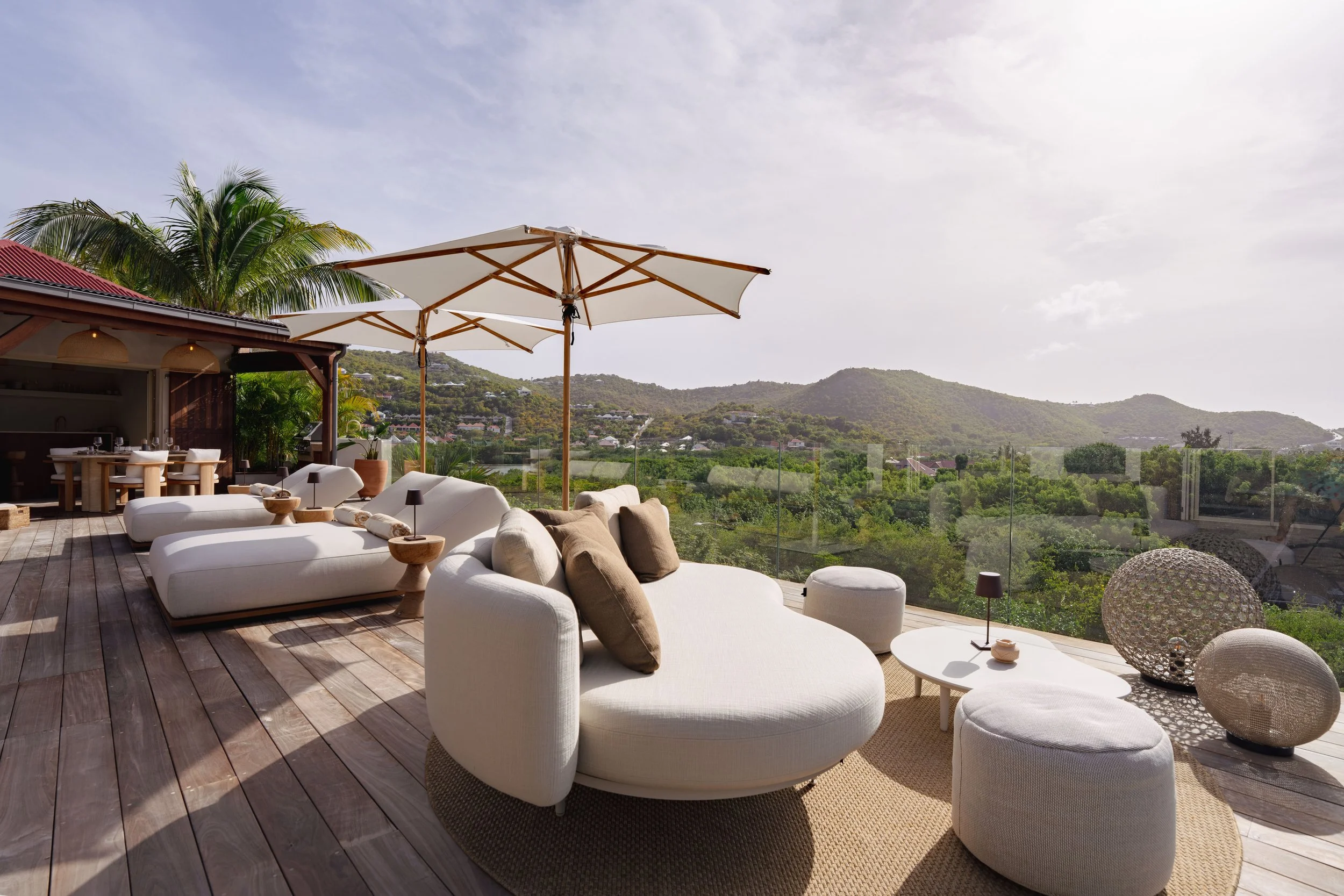 Sunlit outdoor patio with white cushioned seating, beige pillows, a glass railing, lush green hills, and a partly cloudy sky.