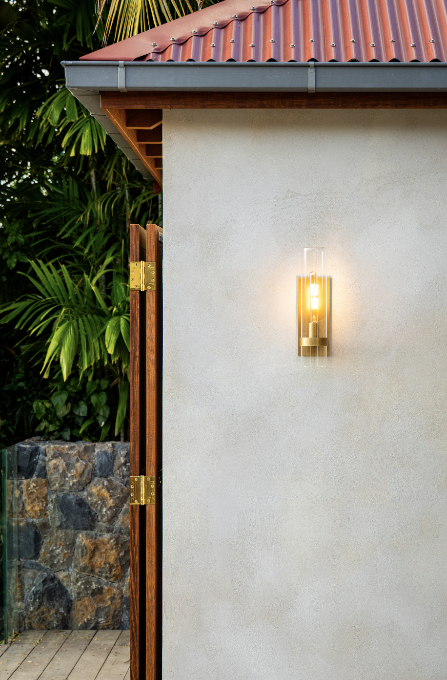 Close-up of a modern house exterior with a wall-mounted light, a partial view of a wooden fence with metal hinges, a red metal roof, and lush green foliage in the background.