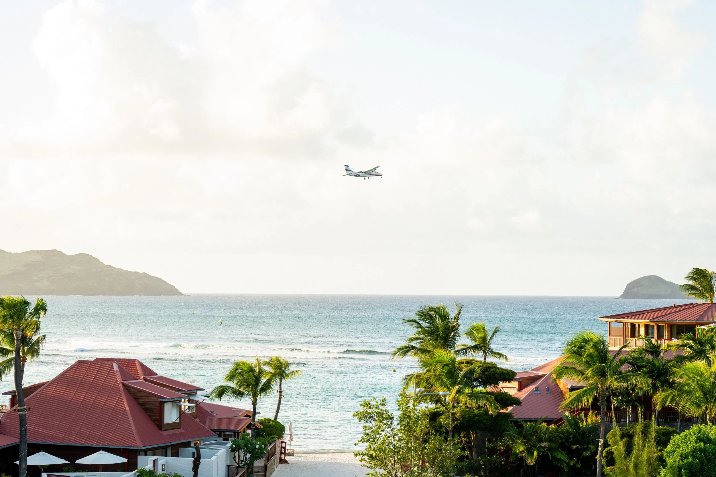 A seaside view with palm trees, houses with red rooftops, ocean water, and distant islands under a partly cloudy sky, with an airplane flying overhead.