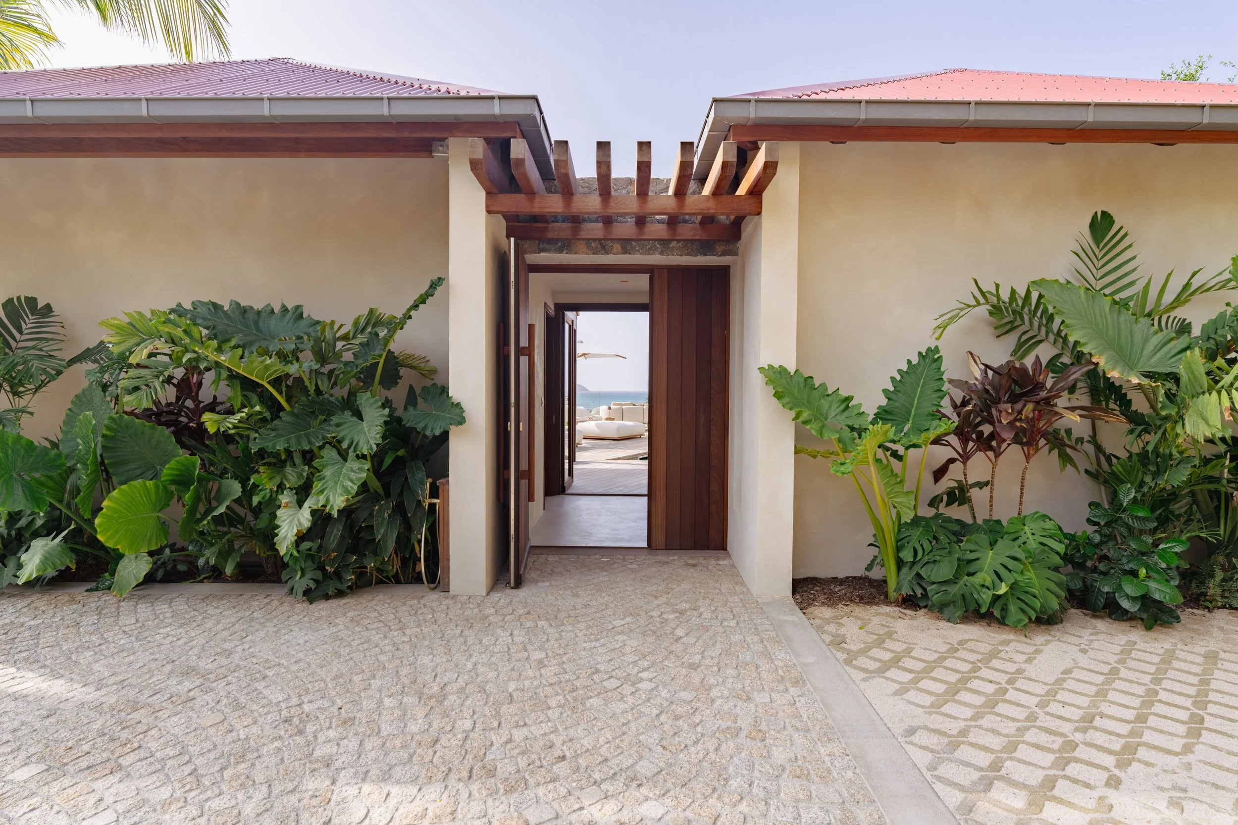 Entrance to a modern house with tropical plants on either side, open door leading to a living room and ocean view, cobblestone pathway.