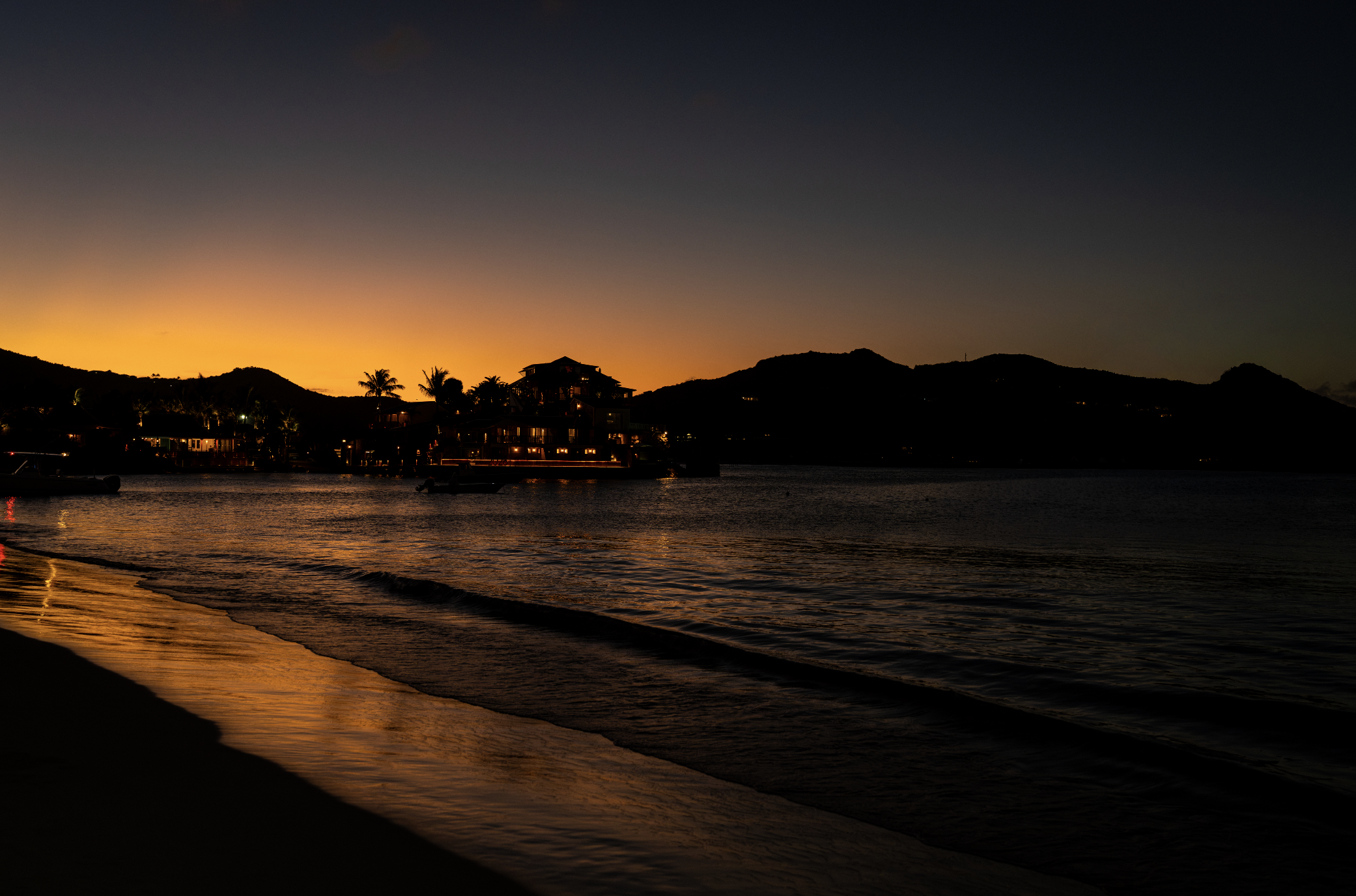 A sunset over a body of water with a silhouette of mountains and houses, with palm trees and boats visible along the shoreline.