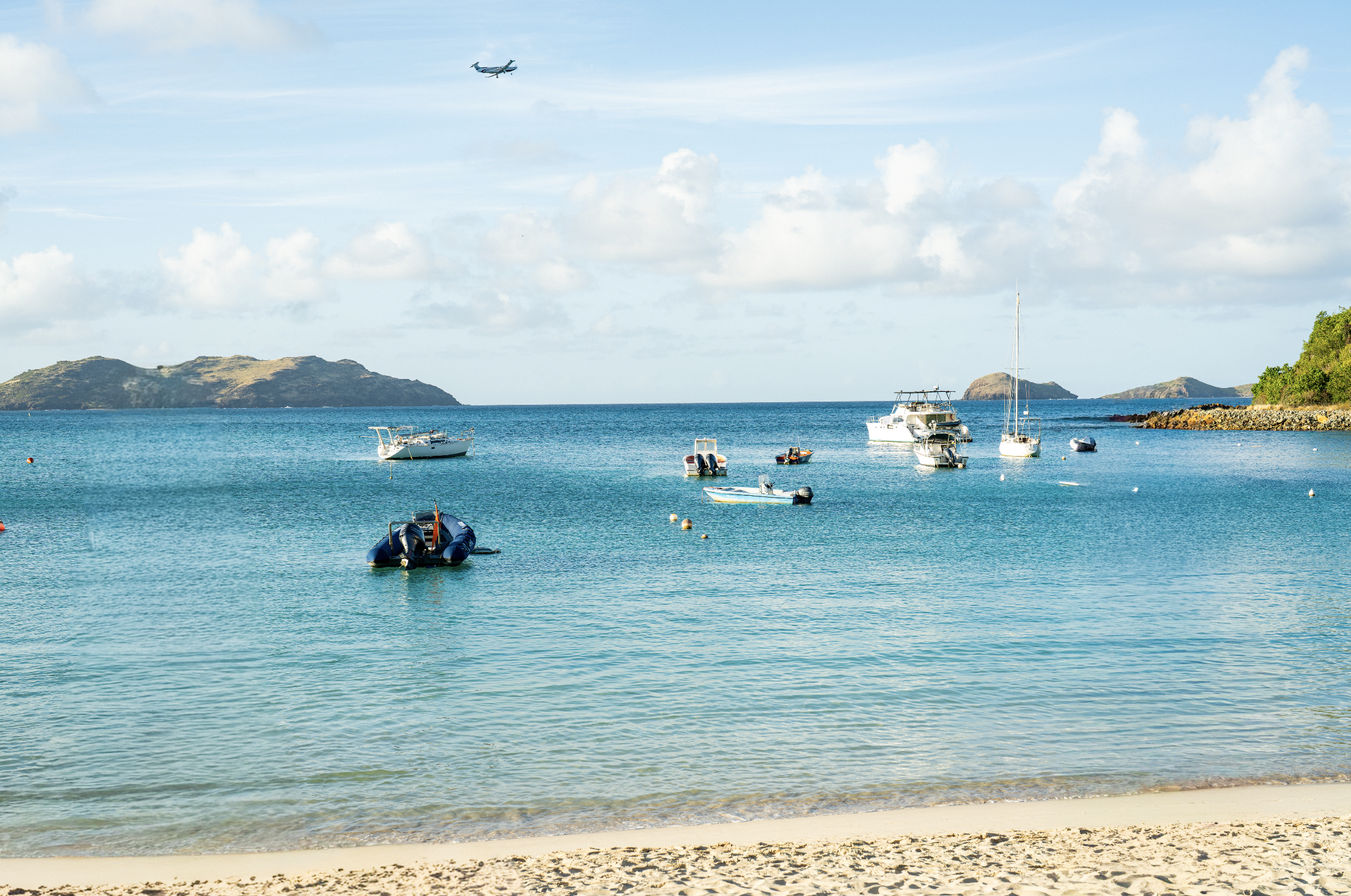 A serene beach scene with clear blue water, small boats anchored near the shore, a boat in the sky, and distant islands under a partly cloudy sky.