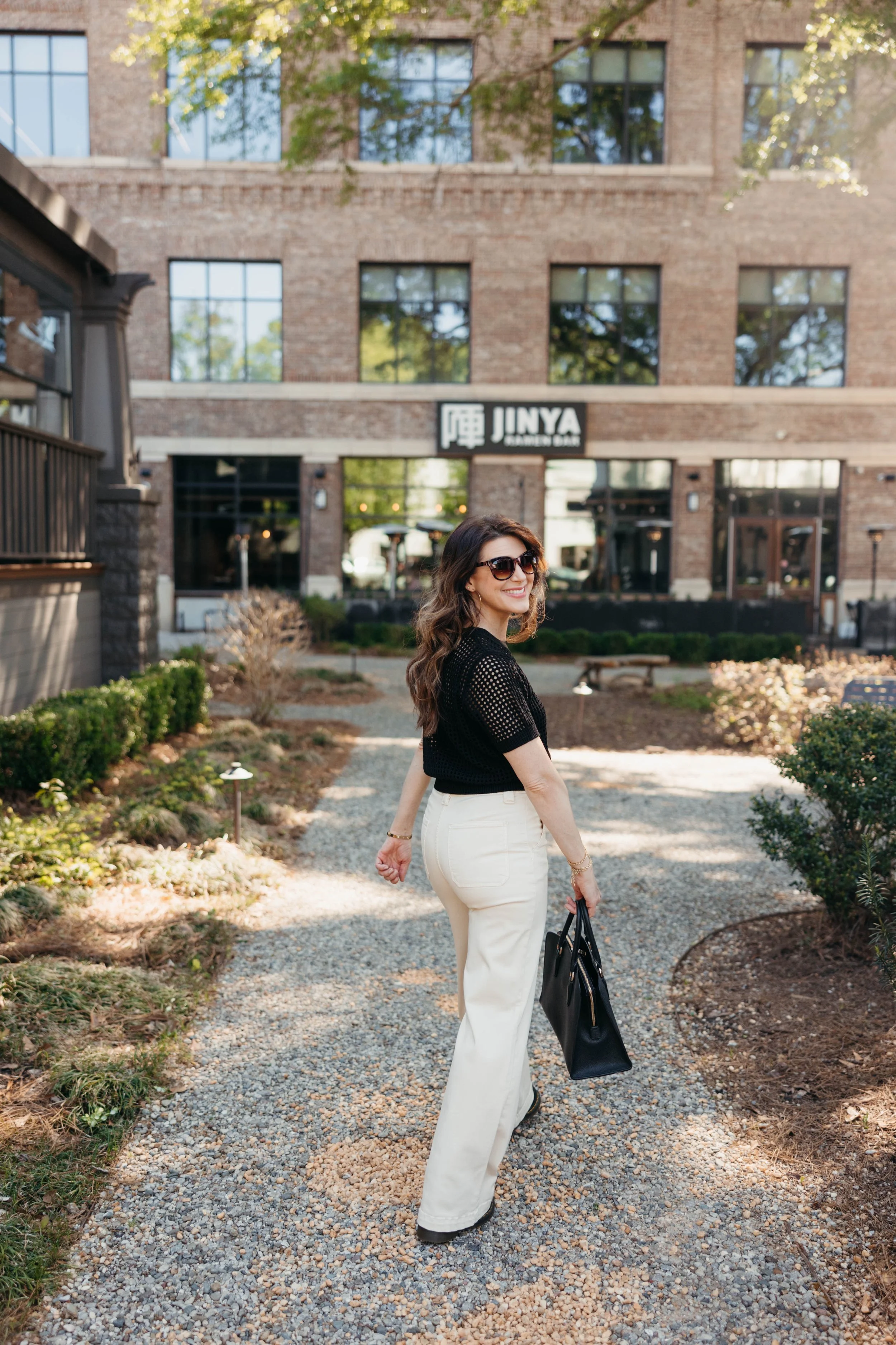 A woman with long wavy hair wearing sunglasses, a black top, and white pants is walking on a gravel path outside of a brick building with large windows. She is holding a black tote bag and smiling.