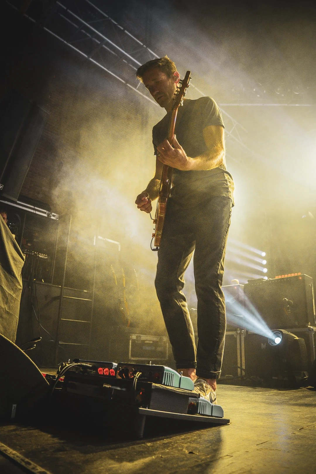 A musician playing an electric guitar on stage, illuminated by stage lights, with effects pedals and equipment in the foreground.
