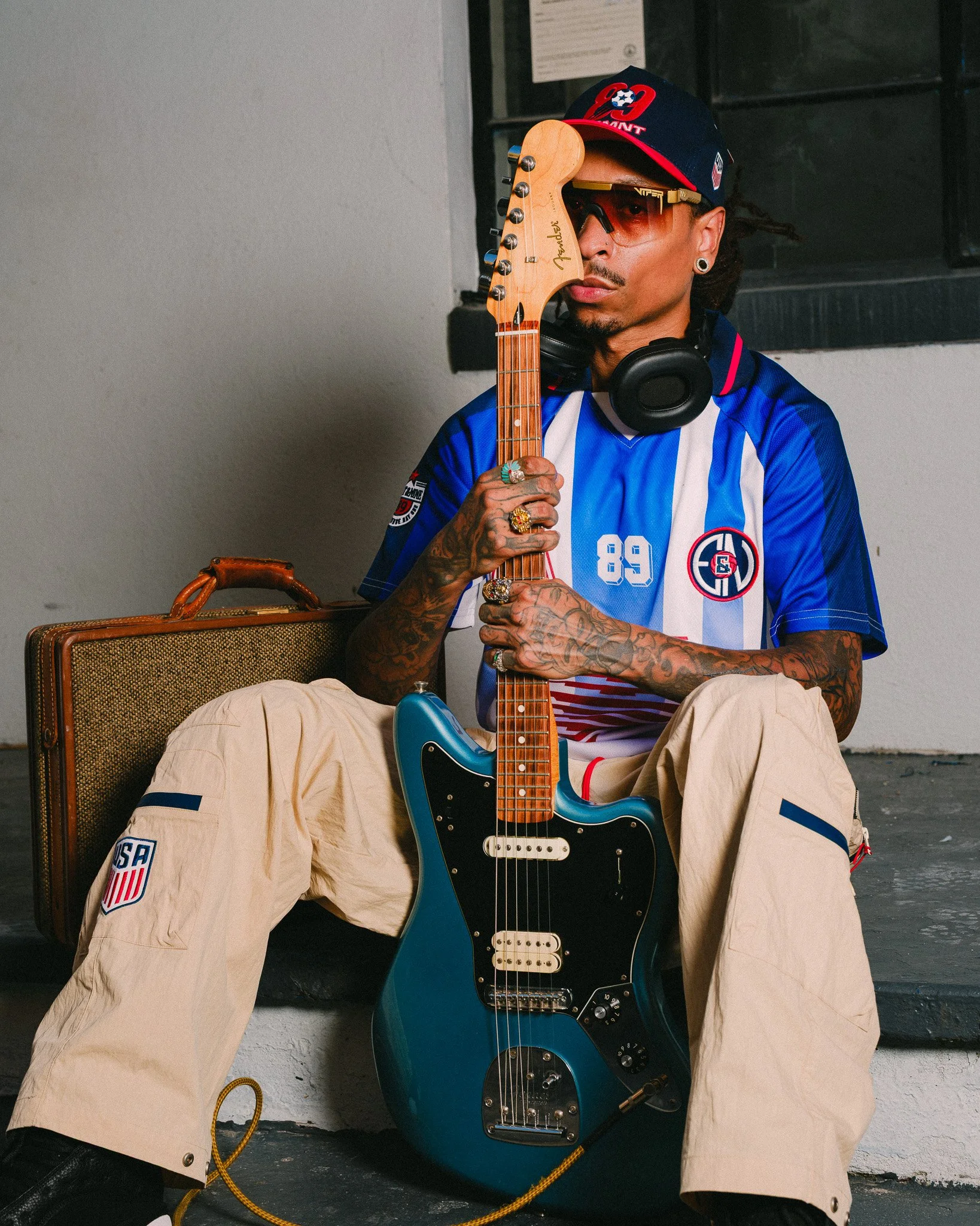 A man wearing a blue Paris Saint-Germain soccer jersey, beige cargo pants with USA patches, a baseball cap, sunglasses, and earrings, sitting against a wall, holding a Fender electric guitar, with black headphones around his neck, and a guitar case n
