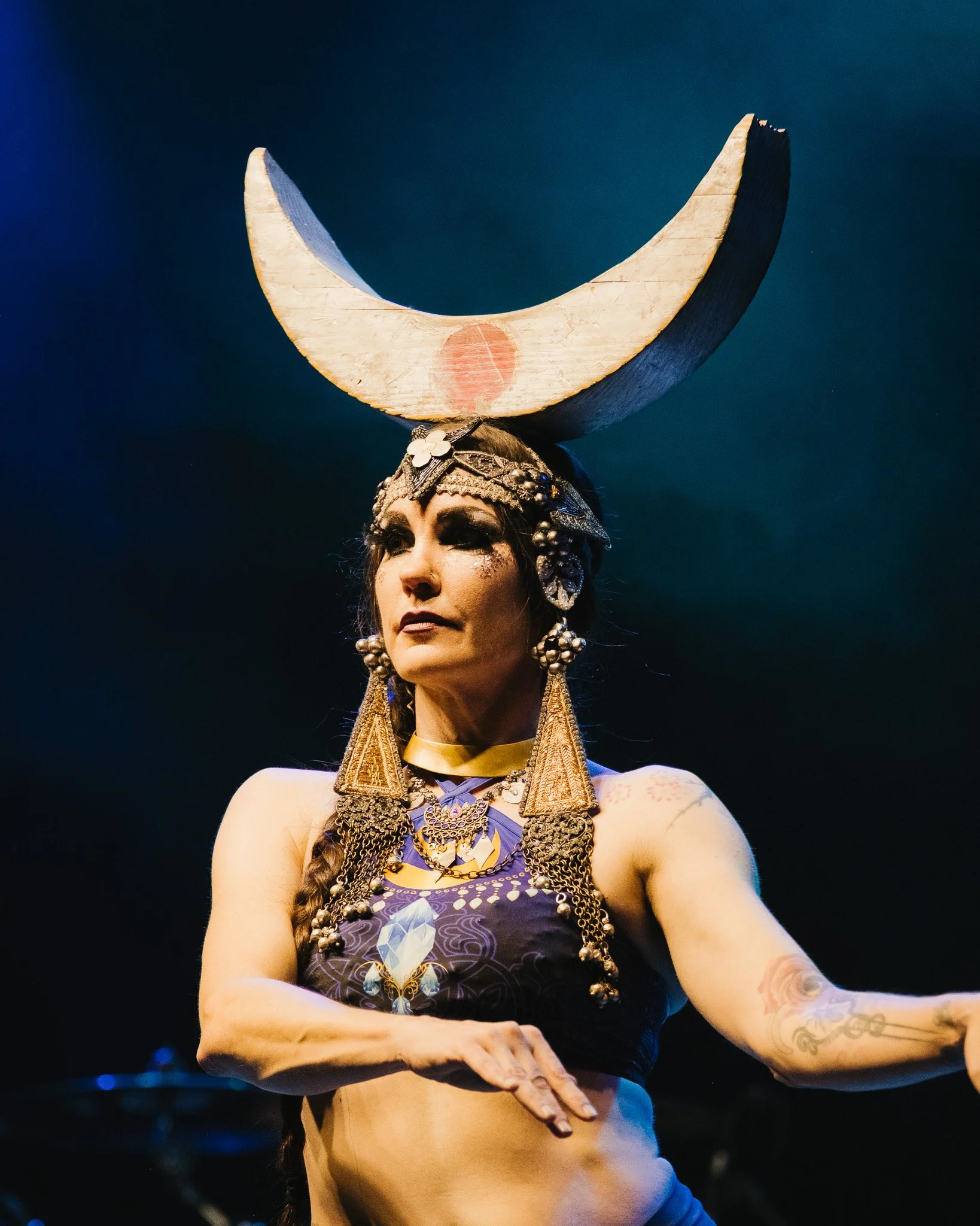 A woman with elaborate jewelry and makeup, wearing a large crescent moon headdress standing against a dark background.