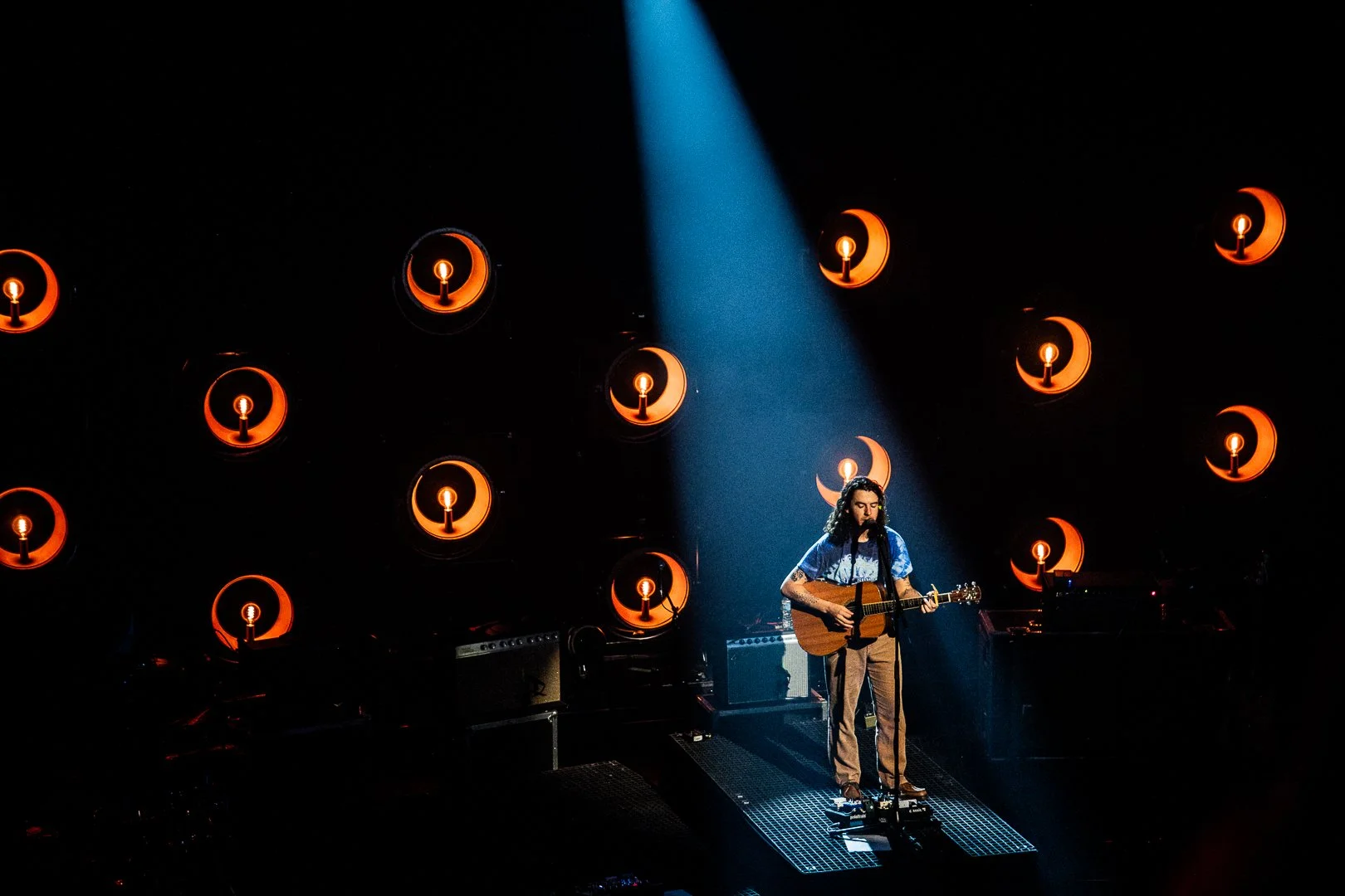 A man with long dark hair and tattoos on his arms is playing an acoustic guitar and singing into a microphone on a dimly lit stage. There is a spotlight focusing on him, and a backdrop of orange lighting fixtures shaped like crescent moons and circle
