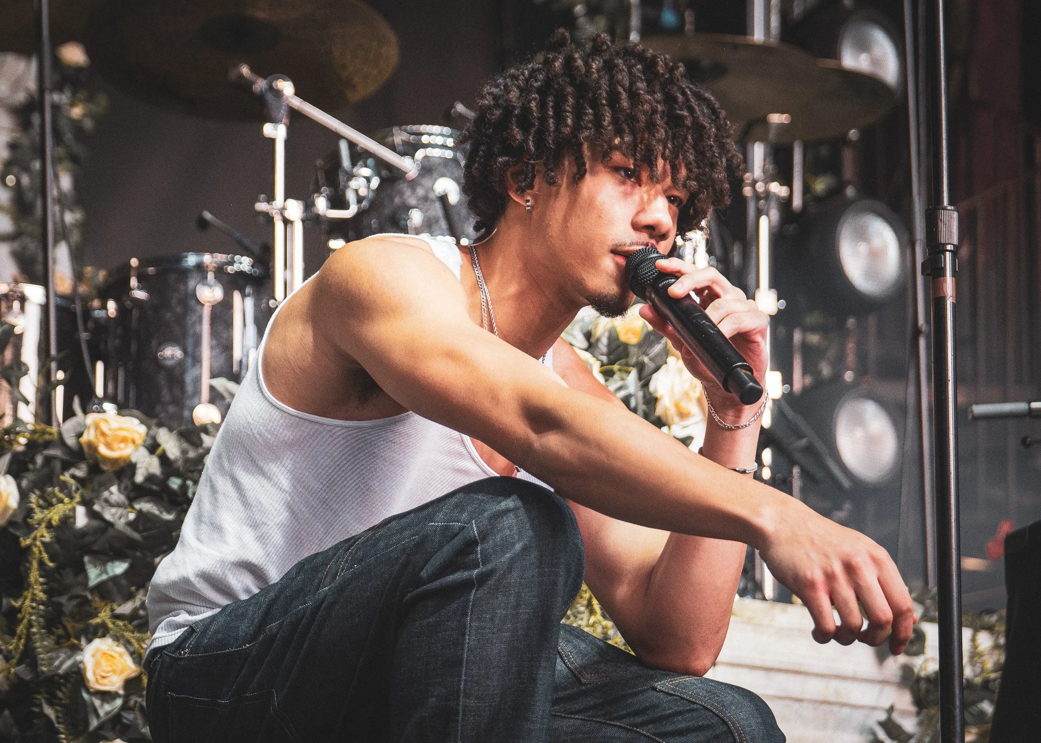 A young man with curly hair and earrings holding a microphone and sitting on the ground in front of a floral arrangement, with speakers and sound equipment in the background.