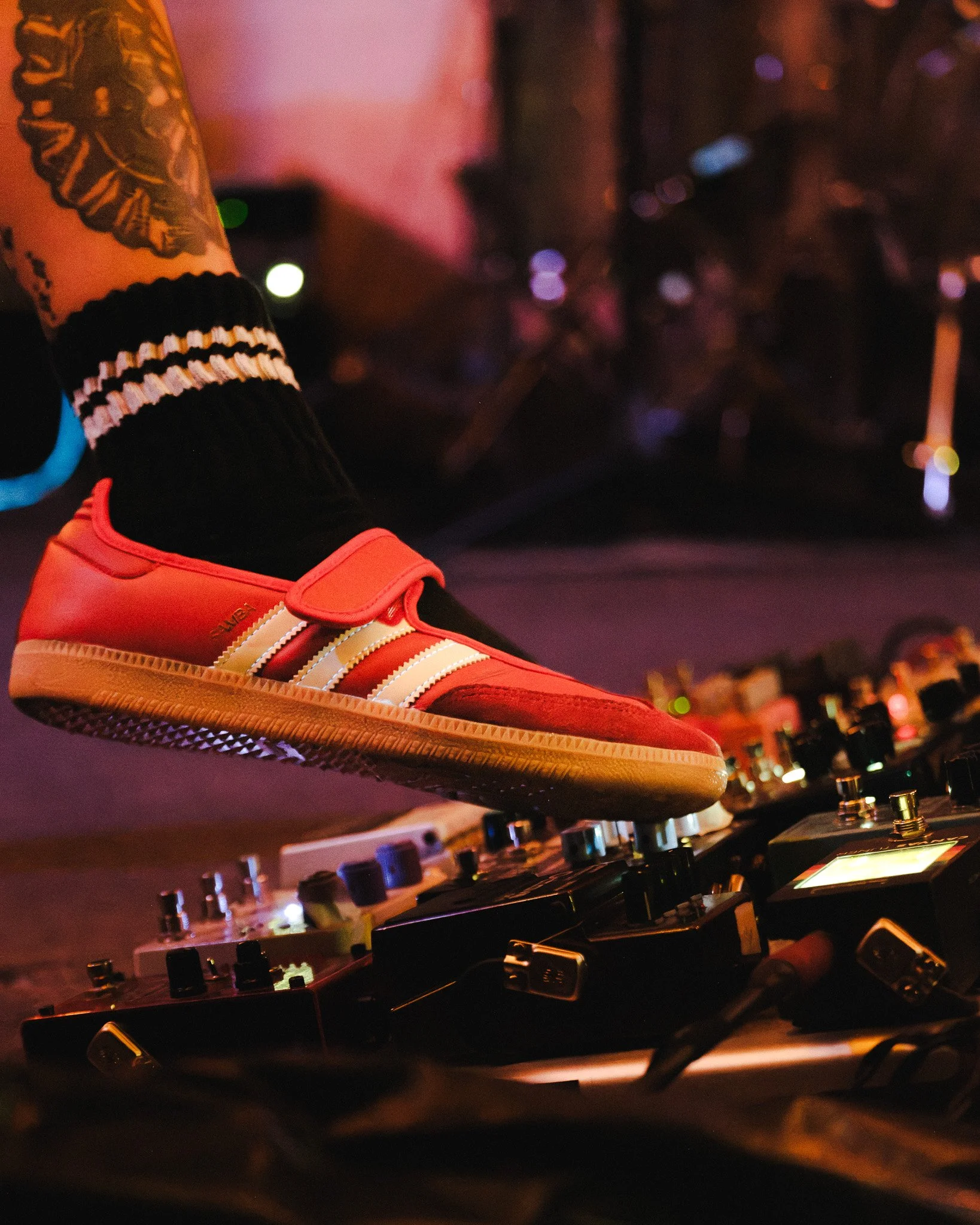 Close-up of a DJ's mixing console and a DJ's foot wearing an orange sneaker, with a background of blurred colorful lights.