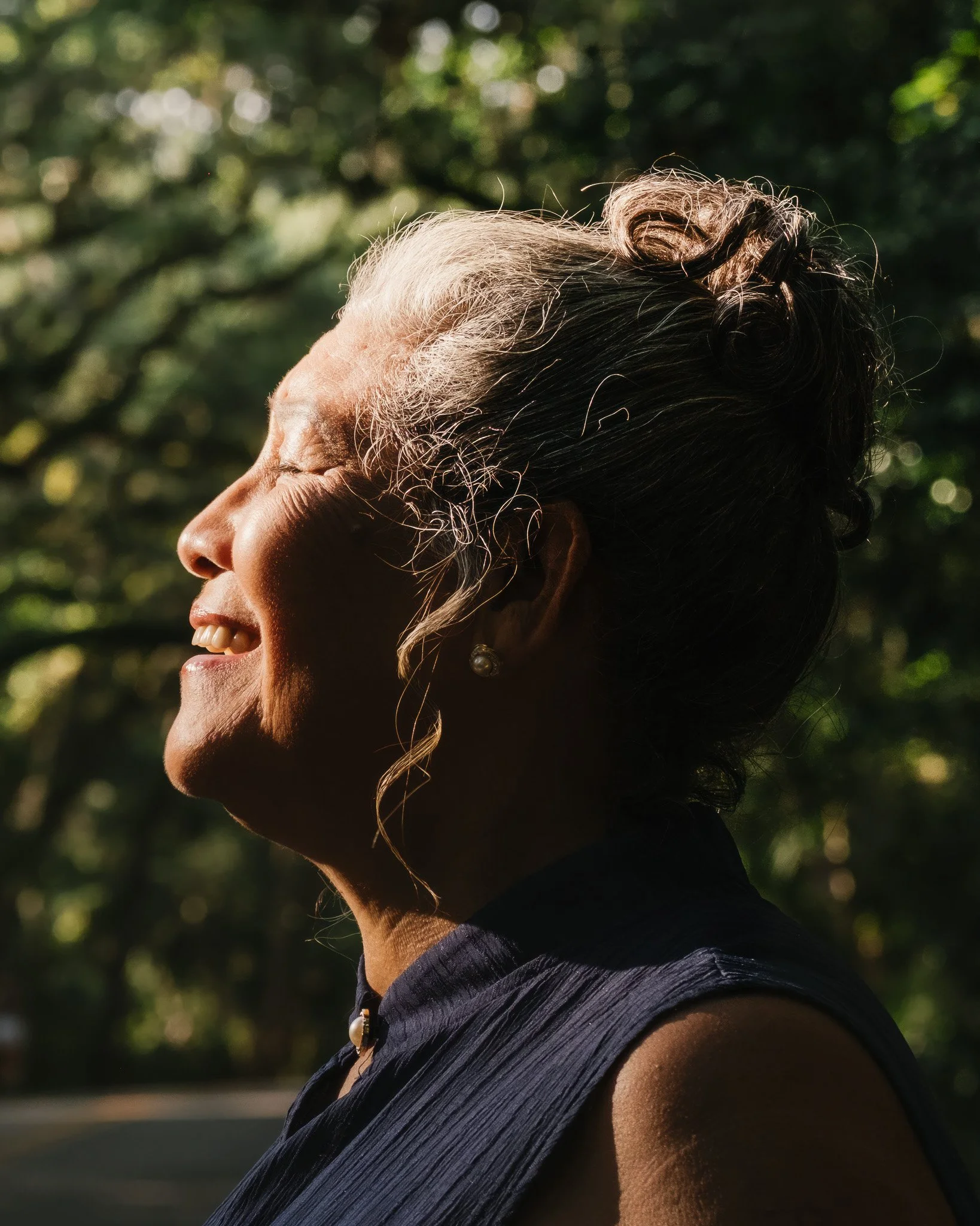 Close-up of an elderly woman with gray hair styled in a bun, smiling with closed eyes, wearing pearl earrings, outdoors with blurred greenery in the background.