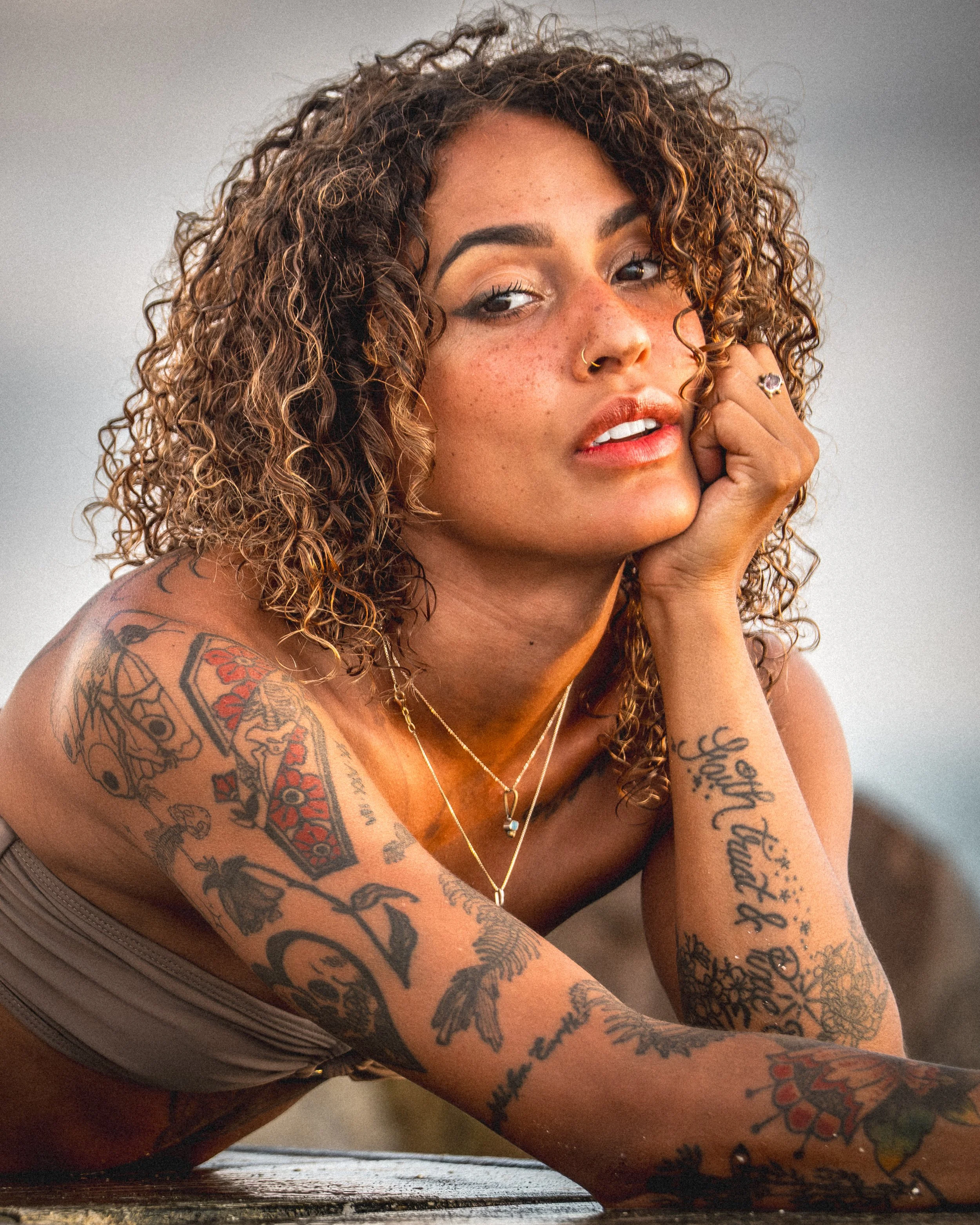A woman with curly brown hair and tattoos on her left arm resting her face on her hand, wearing a grey tank top, and looking directly at the camera.