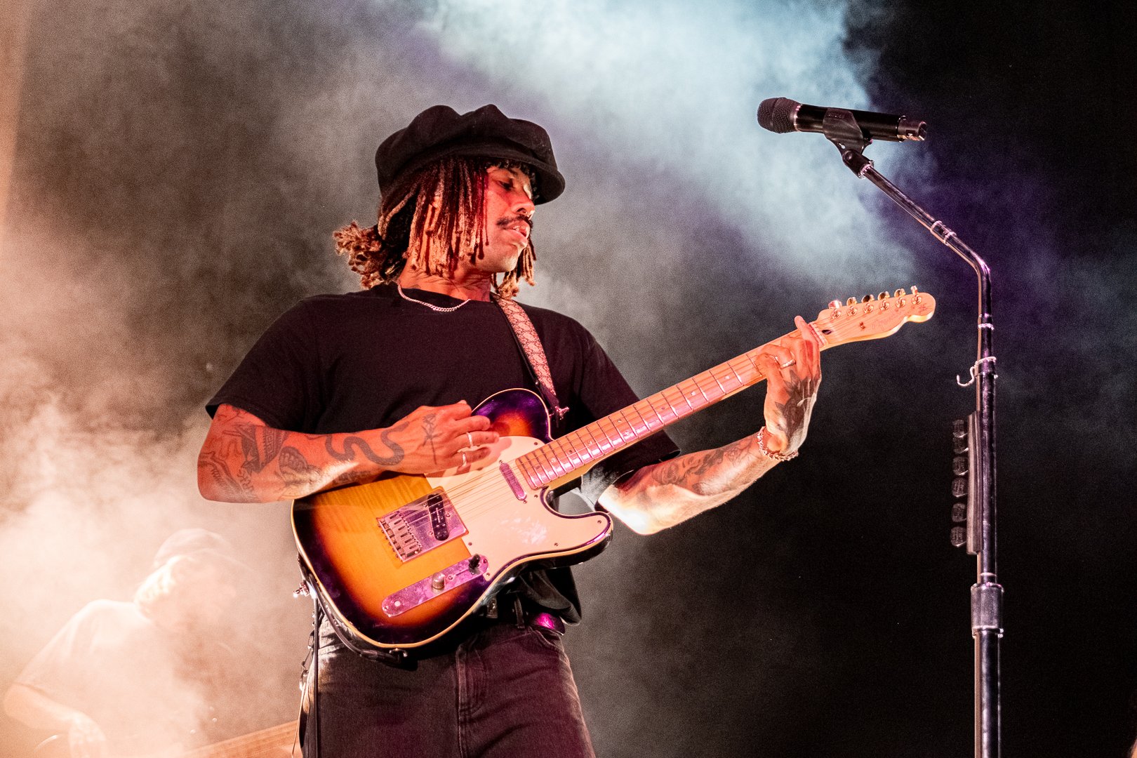 A man with tattoos playing an electric guitar on stage, with smoke and lighting effects, microphone on a stand nearby.