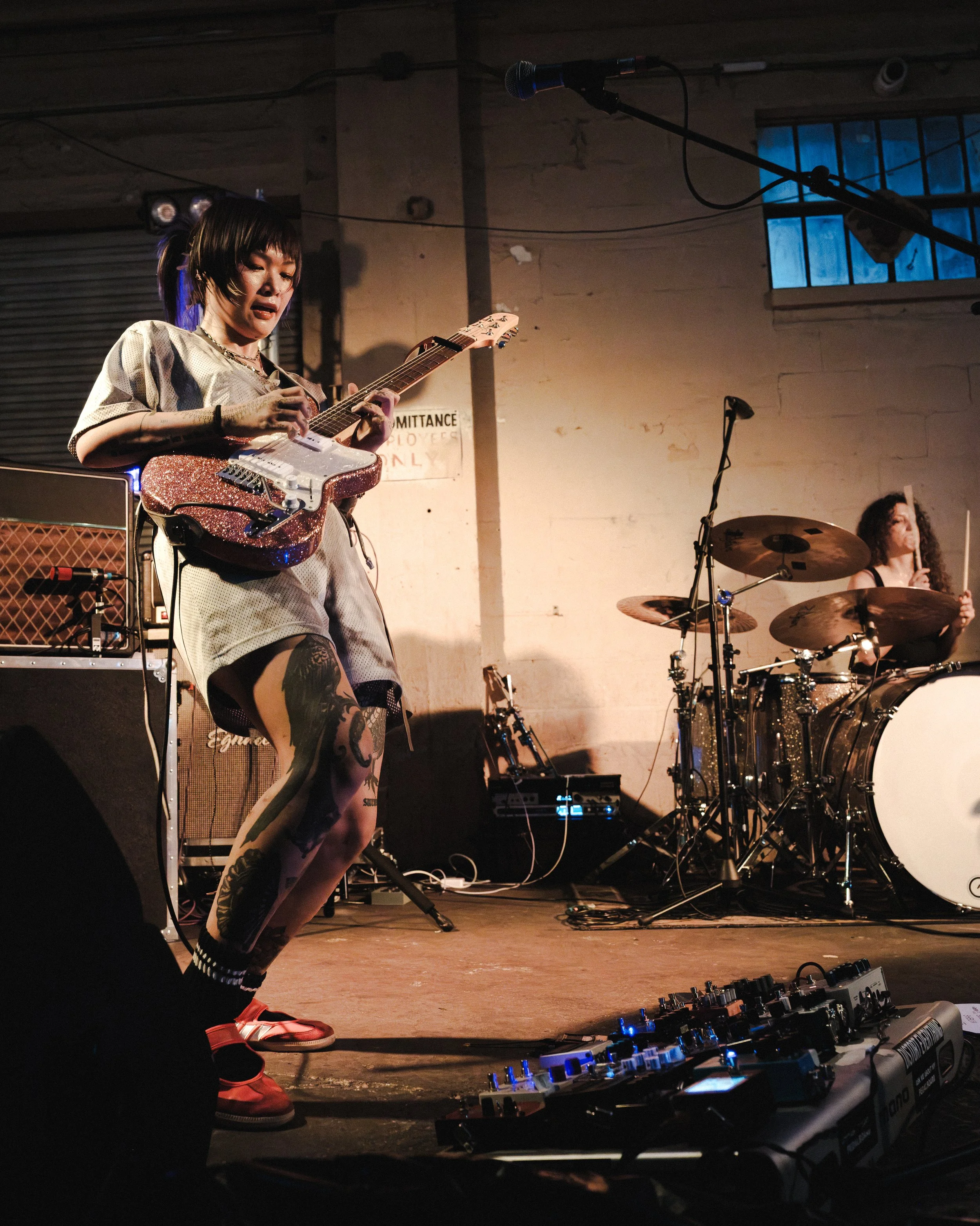 A woman with short dark hair and tattoos on her legs playing an electric guitar on stage. She is wearing casual clothes and red sneakers, and there is a drummer in the background. The stage has musical equipment including a pedalboard and amplifiers,