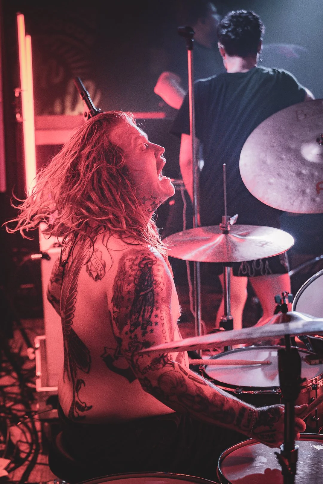 A shirtless man with long, curly hair and numerous tattoos playing a drum set in a dimly lit, pink-colored stage environment.
